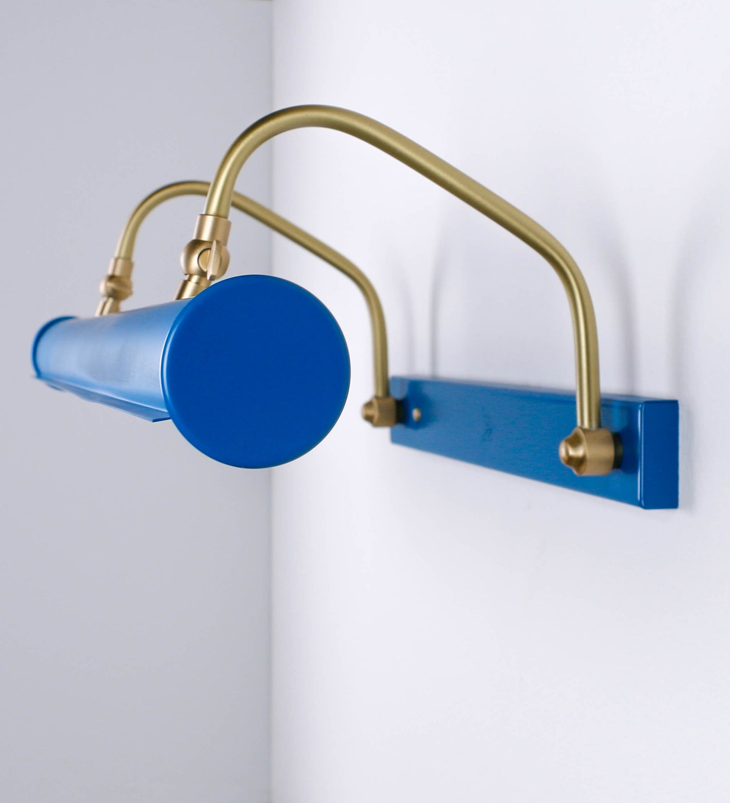 Aplique de parede que combina elementos clássicos com toques contemporâneos. O aplique é composto por uma base retangular azul vintage, de onde se estendem dois braços curvos e graciosos, em latão escovado, que suportam a barra principal. Os braços contêm um elemento, em latão escovado, que permite apertar ou desapertar a barra para melhor manipular a direção da luz. A barra de 65cm de largura, é cilíndrica e alongada, azul vintage. As extremidades do cilindro são planas e do mesmo diâmetro.