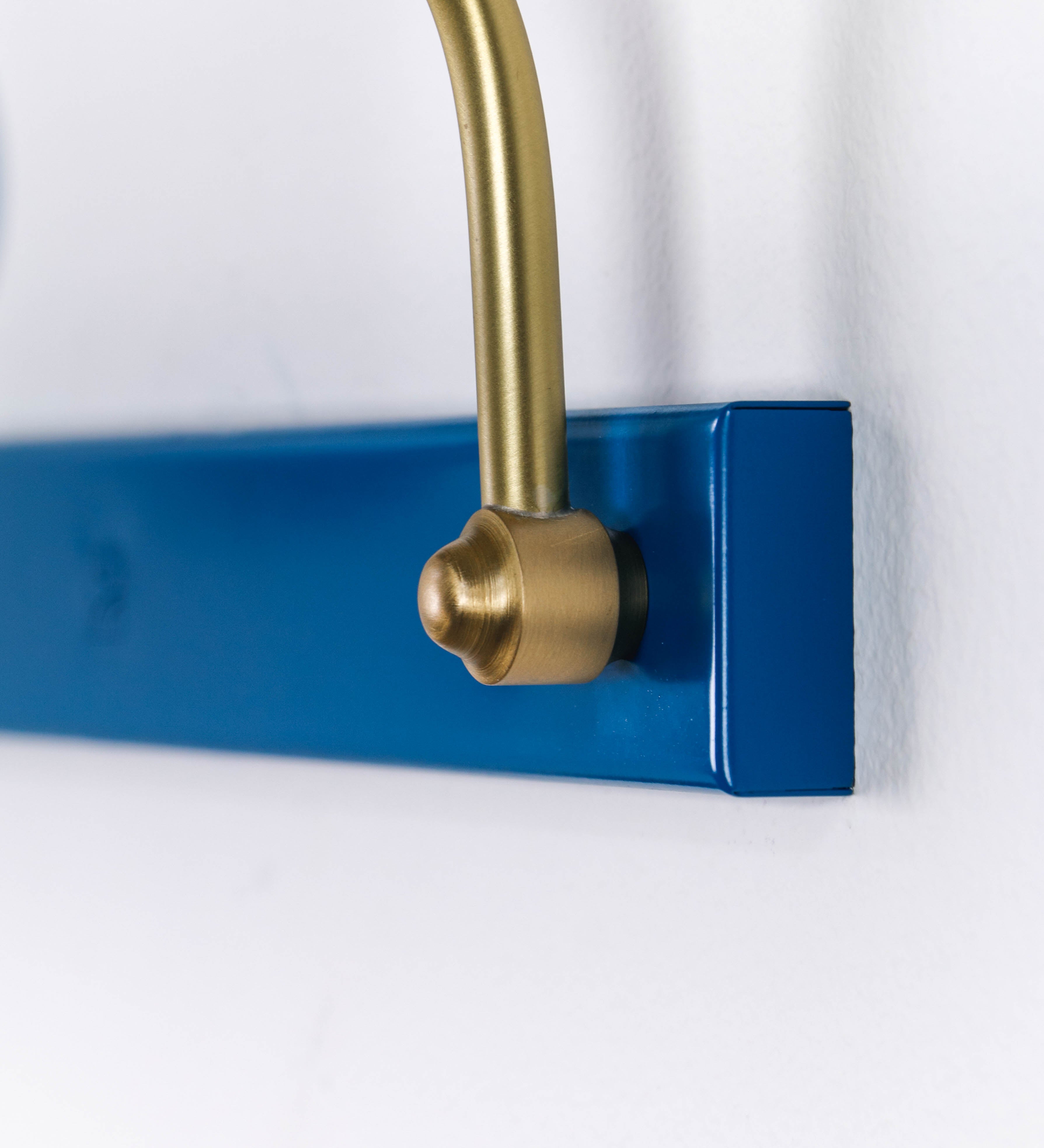 Aplique de parede que combina elementos clássicos com toques contemporâneos. O aplique é composto por uma base retangular azul vintage, de onde se estendem dois braços curvos e graciosos, em latão escovado, que suportam a barra principal. Os braços contêm um elemento, em latão escovado, que permite apertar ou desapertar a barra para melhor manipular a direção da luz. A barra de 65cm de largura, é cilíndrica e alongada, azul vintage. As extremidades do cilindro são planas e do mesmo diâmetro.