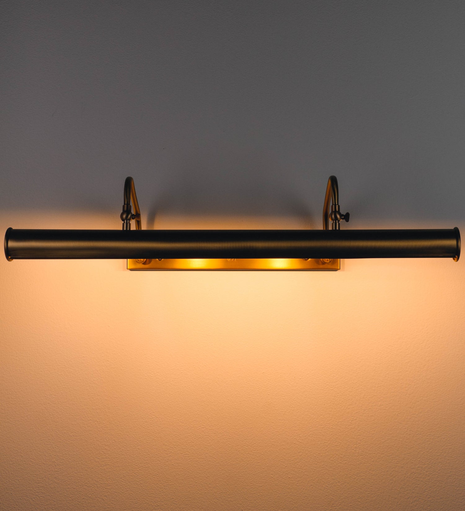 Aplique de parede que combina elementos clássicos com toques contemporâneos, todo em ouro velho. O aplique é composto por uma base retangular, de onde se estendem dois braços curvos e graciosos, que suportam a barra principal. Os braços contêm um elemento que permite apertar ou desapertar a barra para melhor manipular a direção da luz. A barra de 65cm de largura é cilíndrica e alongada. As extremidades do cilindro são planas e do mesmo diâmetro.
