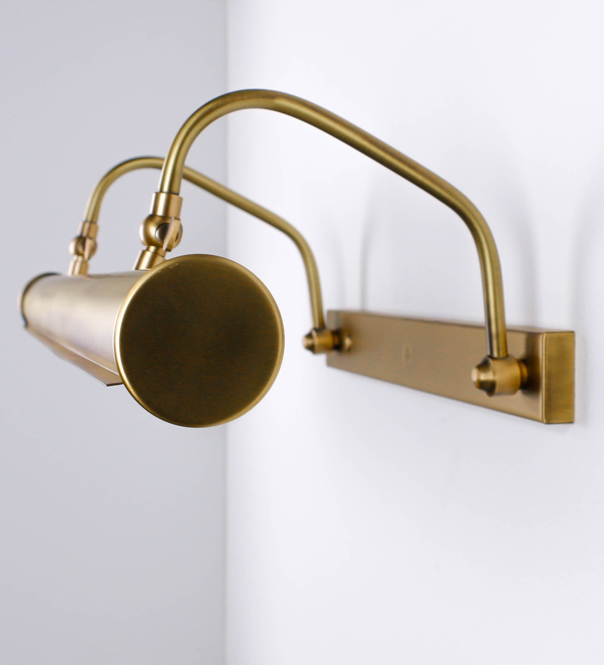 Aplique de parede que combina elementos clássicos com toques contemporâneos, todo em ouro velho. O aplique é composto por uma base retangular, de onde se estendem dois braços curvos e graciosos, que suportam a barra principal. Os braços contêm um elemento que permite apertar ou desapertar a barra para melhor manipular a direção da luz. A barra de 65cm de largura é cilíndrica e alongada. As extremidades do cilindro são planas e do mesmo diâmetro.