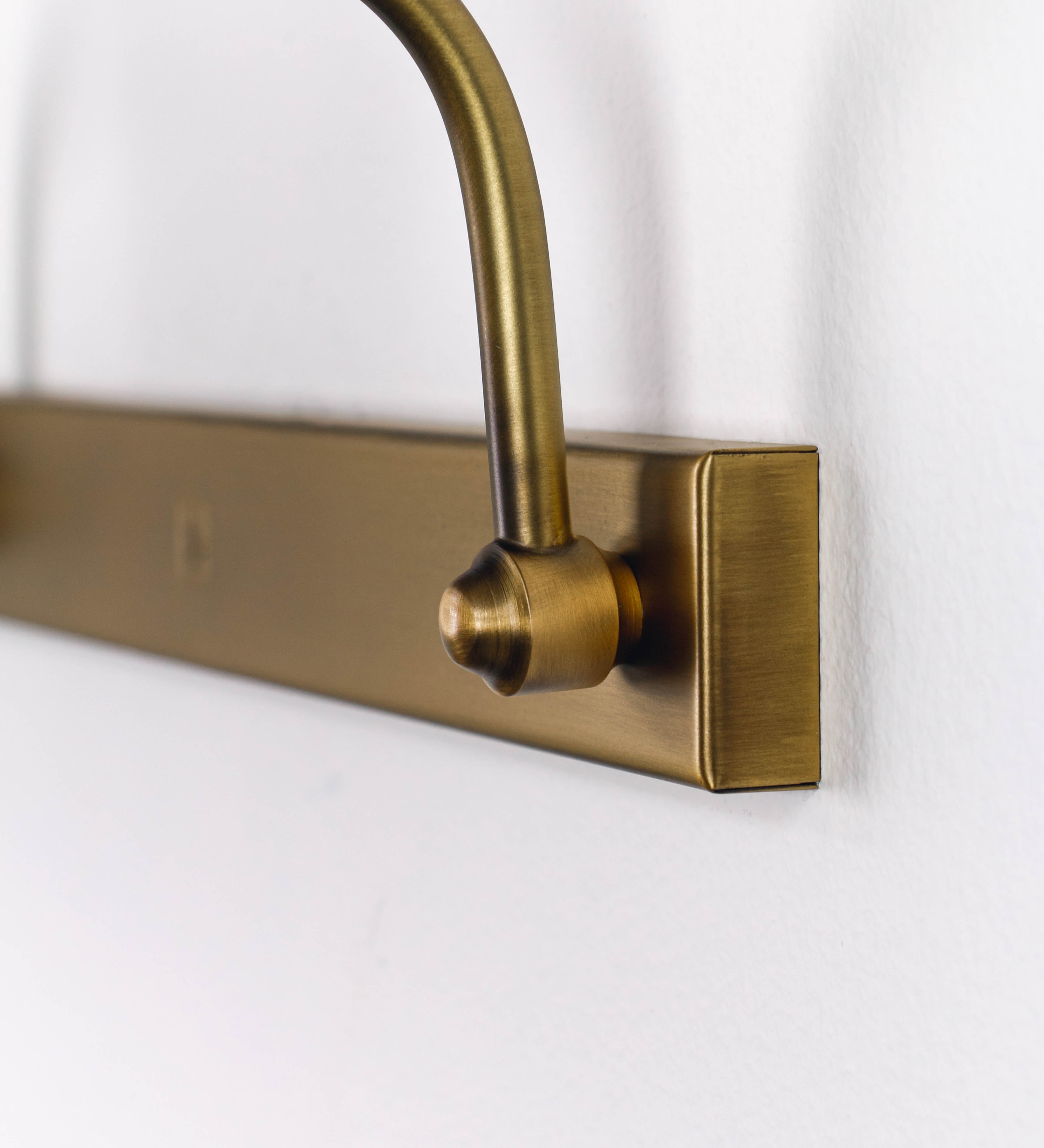 Aplique de parede que combina elementos clássicos com toques contemporâneos, todo em ouro velho. O aplique é composto por uma base retangular, de onde se estendem dois braços curvos e graciosos, que suportam a barra principal. Os braços contêm um elemento que permite apertar ou desapertar a barra para melhor manipular a direção da luz. A barra de 65cm de largura é cilíndrica e alongada. As extremidades do cilindro são planas e do mesmo diâmetro.