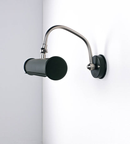 Aplique de parede que combina elementos clássicos com toques contemporâneos. O aplique é composto por uma base circular verde-azeitona, de onde se estende um braço curvo e gracioso, em níquel escovado, que suporta a barra principal. O braço contém um elemento, em níquel escovado, que permite apertar ou desapertar a barra para melhor manipular a direção da luz. A barra de 25cm de largura, é cilíndrica e alongada, verde-azeitona. As extremidades do cilindro são planas e do mesmo diâmetro.