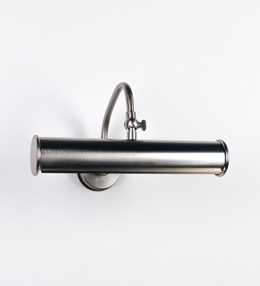 Aplique de parede que combina elementos clássicos com toques contemporâneos, todo em níquel escovado. O aplique é composto por uma base circular de onde se estende um braço curvo e gracioso, que suporta a barra principal. O braço contém um elemento que permite apertar ou desapertar a barra para melhor manipular a direção da luz. A barra de 25cm de largura é cilíndrica e alongada. As extremidades do cilindro são planas e do mesmo diâmetro.