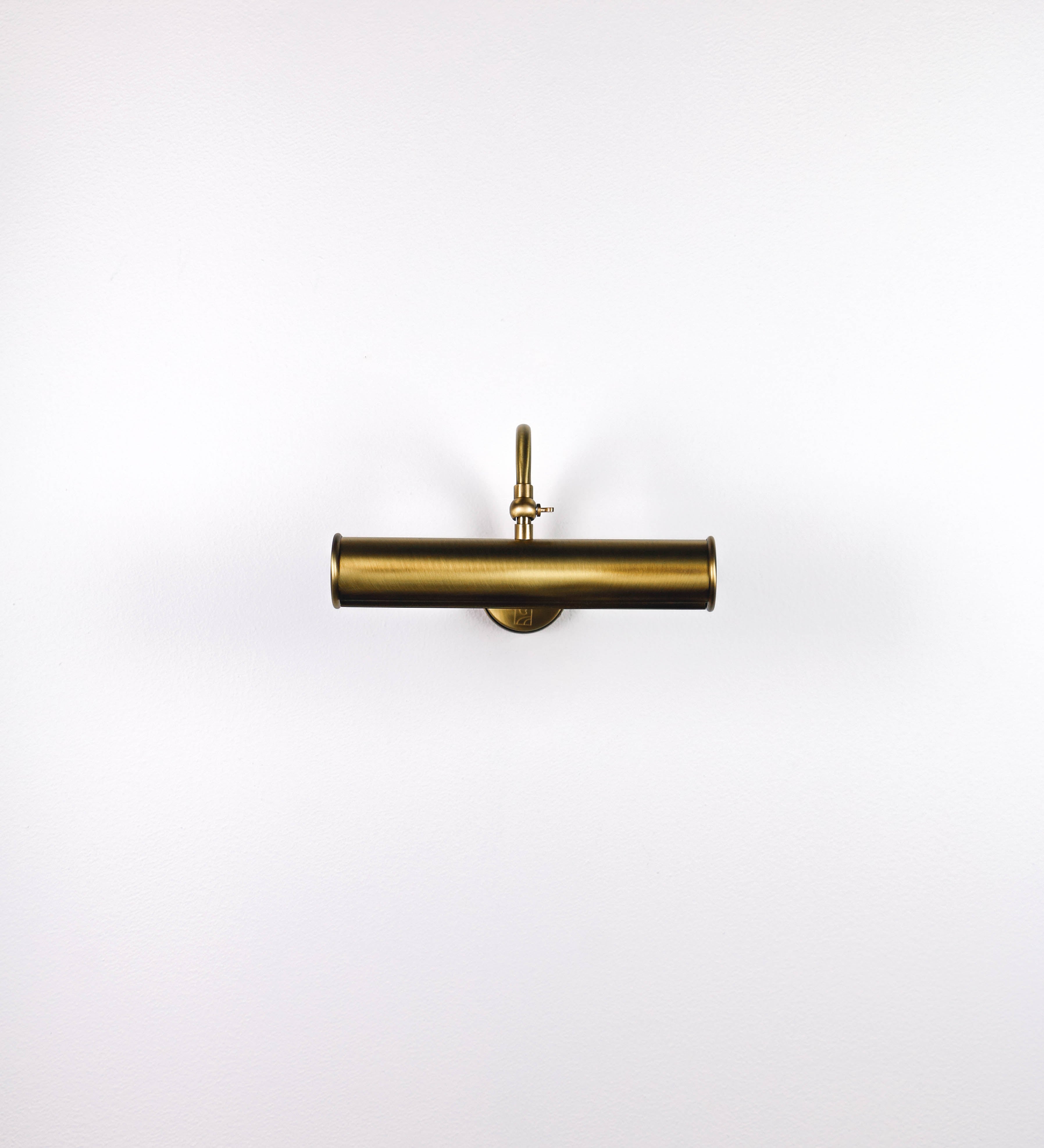 Aplique de parede que combina elementos clássicos com toques contemporâneos, todo em ouro velho. O aplique é composto por uma base circular de onde se estende um braço curvo e gracioso, que suporta a barra principal. O braço contém um elemento que permite apertar ou desapertar a barra para melhor manipular a direção da luz. A barra de 25cm de largura é cilíndrica e alongada. As extremidades do cilindro são planas e do mesmo diâmetro.
