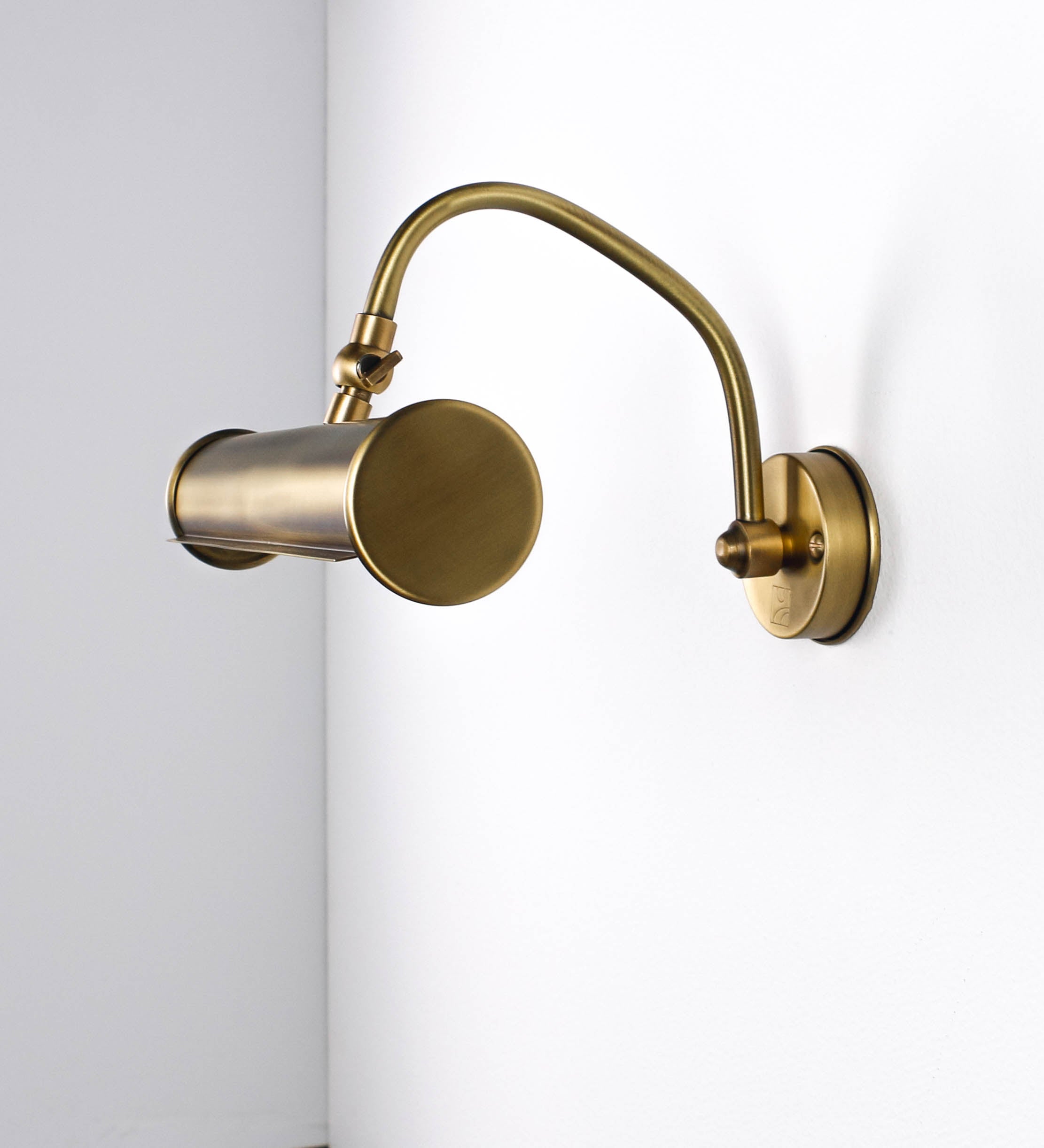 Aplique de parede que combina elementos clássicos com toques contemporâneos, todo em ouro velho. O aplique é composto por uma base circular de onde se estende um braço curvo e gracioso, que suporta a barra principal. O braço contém um elemento que permite apertar ou desapertar a barra para melhor manipular a direção da luz. A barra de 25cm de largura é cilíndrica e alongada. As extremidades do cilindro são planas e do mesmo diâmetro.