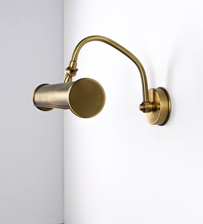 Aplique de parede que combina elementos clássicos com toques contemporâneos, todo em ouro velho. O aplique é composto por uma base circular de onde se estende um braço curvo e gracioso, que suporta a barra principal. O braço contém um elemento que permite apertar ou desapertar a barra para melhor manipular a direção da luz. A barra de 25cm de largura é cilíndrica e alongada. As extremidades do cilindro são planas e do mesmo diâmetro.