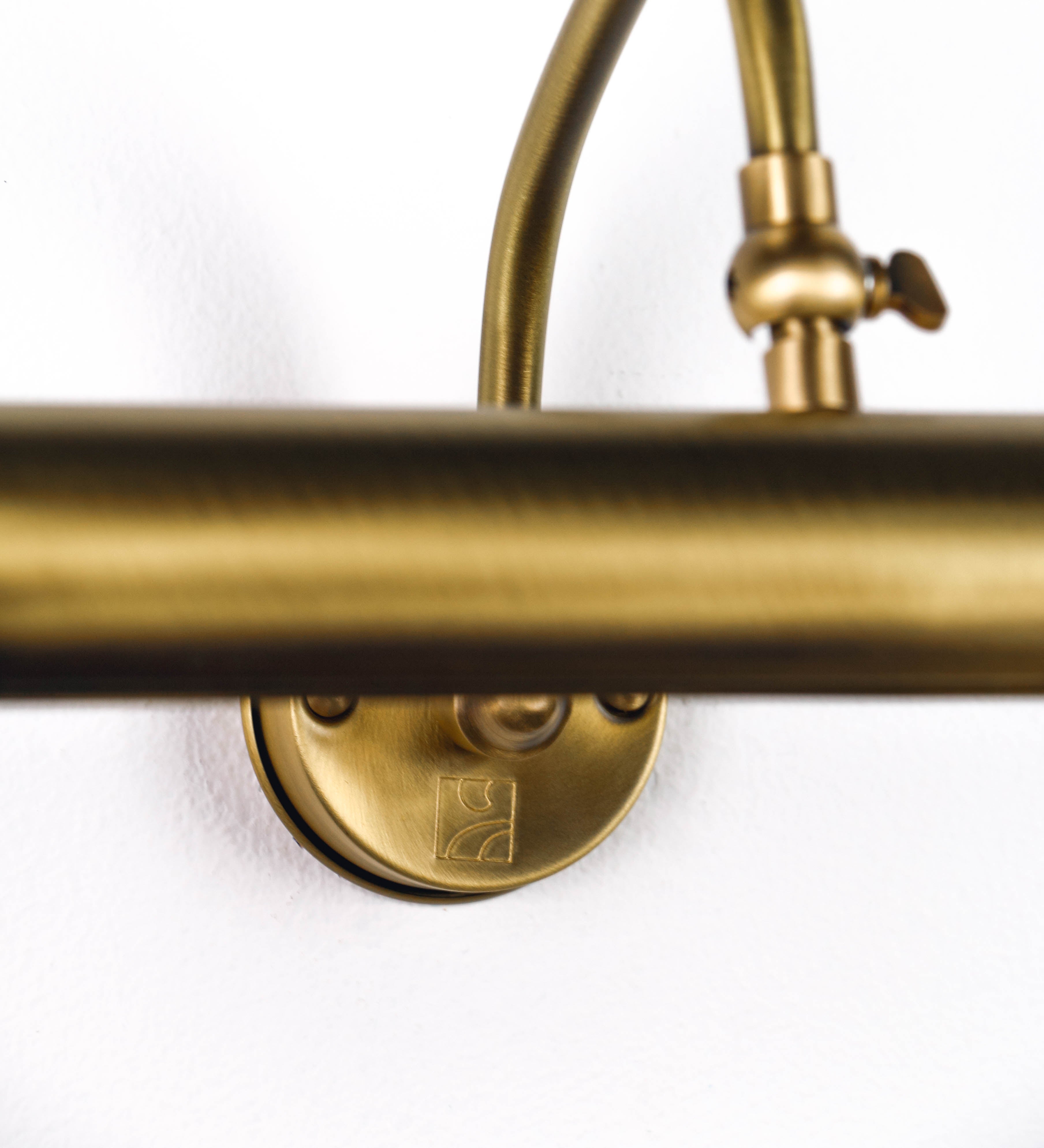 Aplique de parede que combina elementos clássicos com toques contemporâneos, todo em ouro velho. O aplique é composto por uma base circular de onde se estende um braço curvo e gracioso, que suporta a barra principal. O braço contém um elemento que permite apertar ou desapertar a barra para melhor manipular a direção da luz. A barra de 25cm de largura é cilíndrica e alongada. As extremidades do cilindro são planas e do mesmo diâmetro.