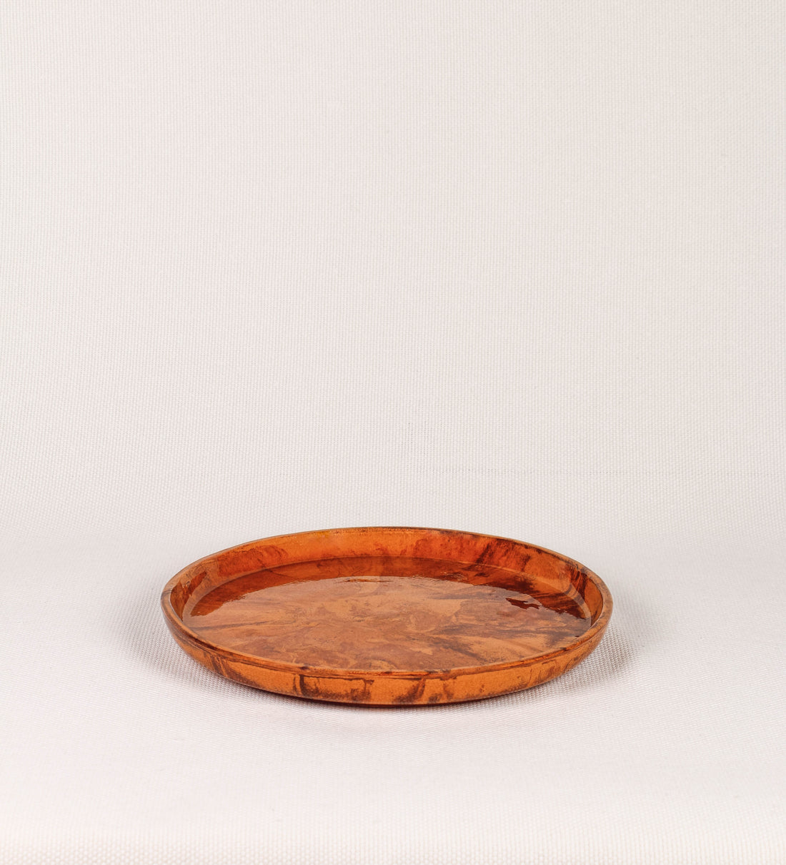 Prato raso com design minimalista e distinto. O prato é de barro, com uma mistura de dois tipos de barro diferentes, um mais escuro e outro mais claro. Esta mistura cria um desenho de formas abstratas e aleatórias, que se espalham por todas as superfícies do prato. Tem acabamento vidrado castanho translúcido, escurecendo as cores originais dos barros misturados. A forma do prato é circular, com uma borda envolvente de altura baixa e design simples e minimalista. 