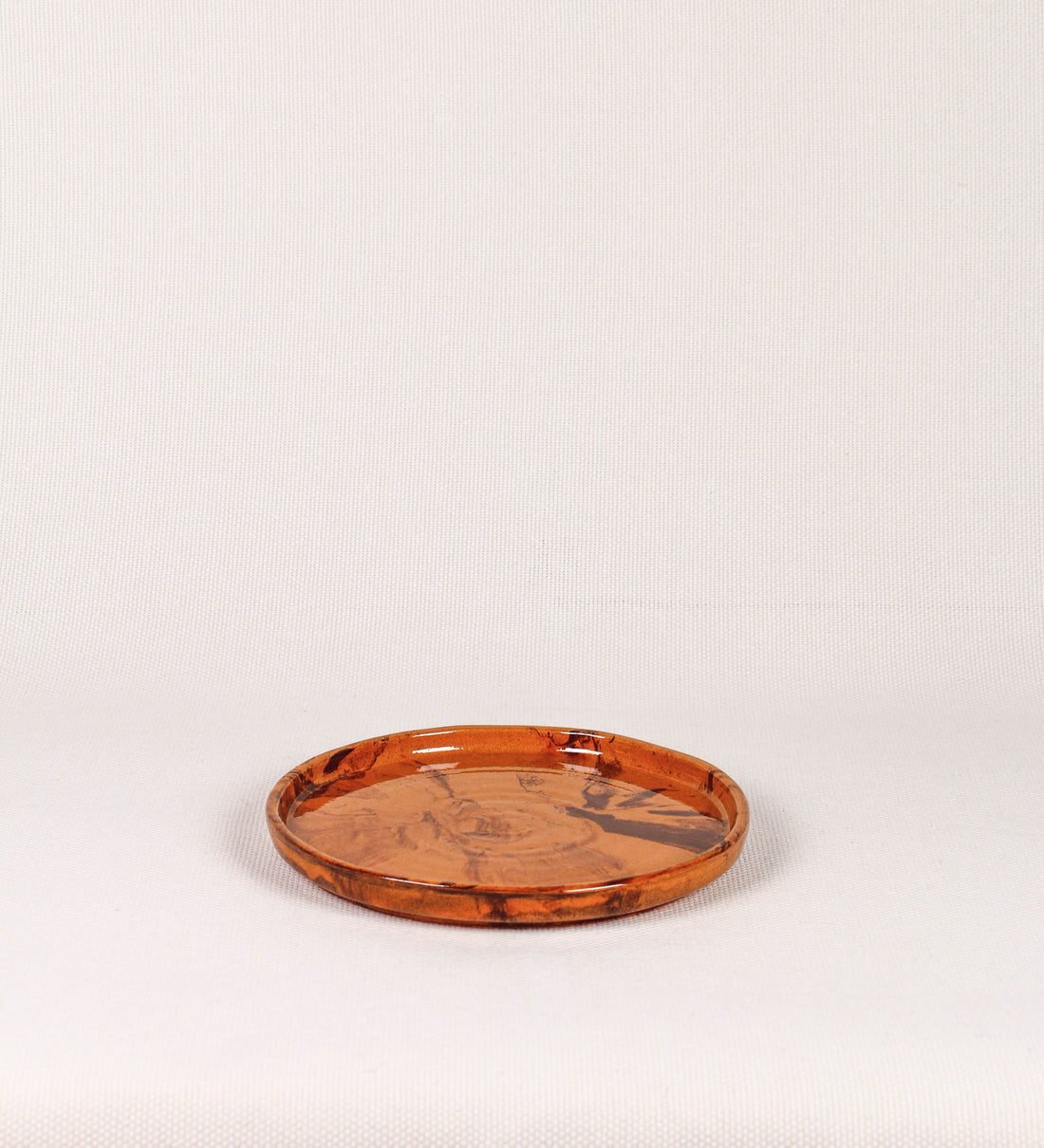 Prato de aperitivos com design minimalista e distinto. O prato é de barro, com uma mistura de dois tipos de barro diferentes, um mais escuro e outro mais claro. Esta mistura cria um desenho de formas abstratas e aleatórias, que se espalham por todas as superfícies do prato. Tem acabamento vidrado castanho translúcido, escurecendo as cores originais dos barros misturados. A forma do prato é circular, com uma borda envolvente de altura baixa e design simples e minimalista. 