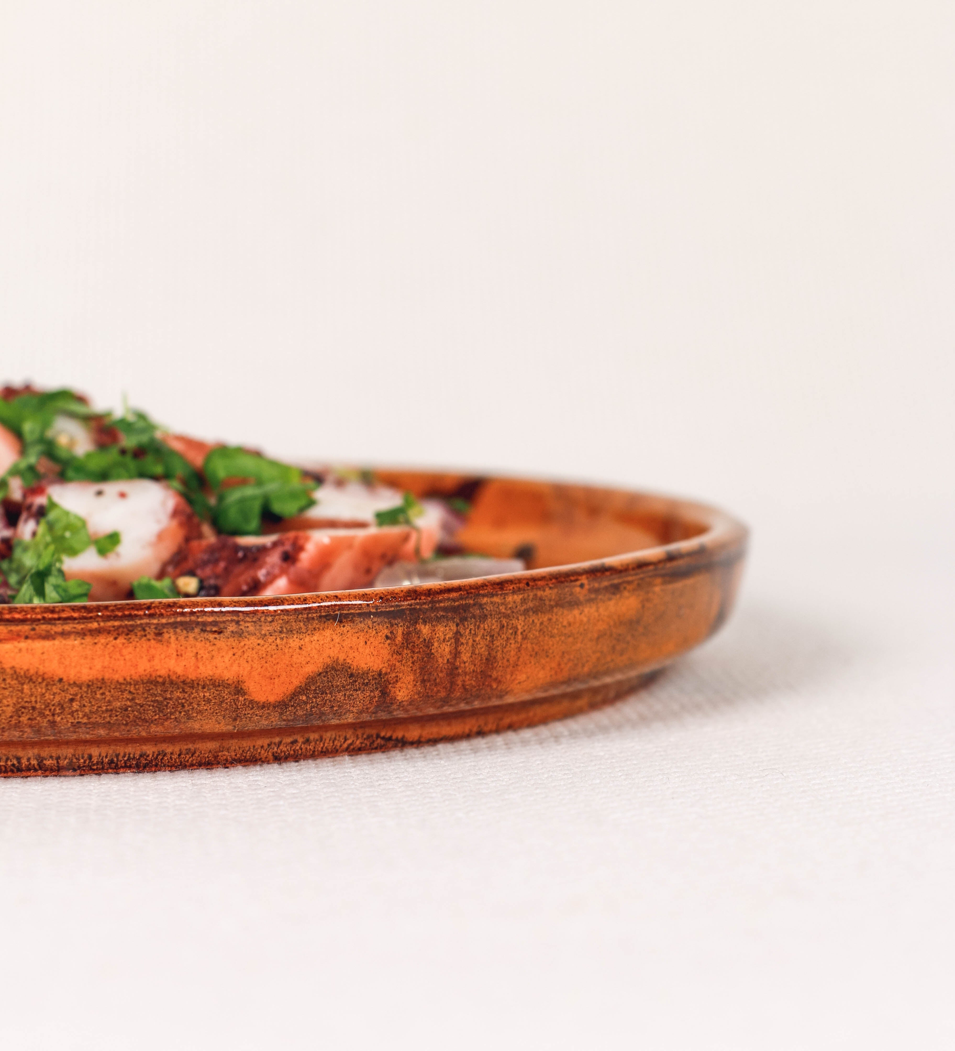 Prato de aperitivos com design minimalista e distinto. O prato é de barro, com uma mistura de dois tipos de barro diferentes, um mais escuro e outro mais claro. Esta mistura cria um desenho de formas abstratas e aleatórias, que se espalham por todas as superfícies do prato. Tem acabamento vidrado castanho translúcido, escurecendo as cores originais dos barros misturados. A forma do prato é circular, com uma borda envolvente de altura baixa e design simples e minimalista. 