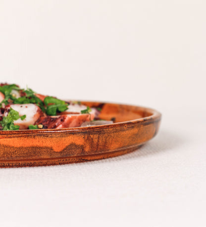 Prato de aperitivos com design minimalista e distinto. O prato é de barro, com uma mistura de dois tipos de barro diferentes, um mais escuro e outro mais claro. Esta mistura cria um desenho de formas abstratas e aleatórias, que se espalham por todas as superfícies do prato. Tem acabamento vidrado castanho translúcido, escurecendo as cores originais dos barros misturados. A forma do prato é circular, com uma borda envolvente de altura baixa e design simples e minimalista. 