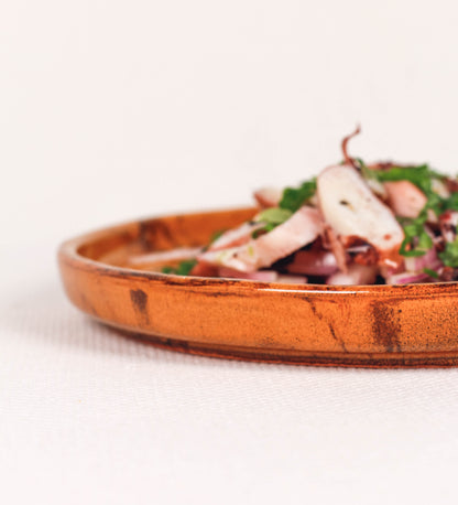 Prato de aperitivos com design minimalista e distinto. O prato é de barro, com uma mistura de dois tipos de barro diferentes, um mais escuro e outro mais claro. Esta mistura cria um desenho de formas abstratas e aleatórias, que se espalham por todas as superfícies do prato. Tem acabamento vidrado castanho translúcido, escurecendo as cores originais dos barros misturados. A forma do prato é circular, com uma borda envolvente de altura baixa e design simples e minimalista. 