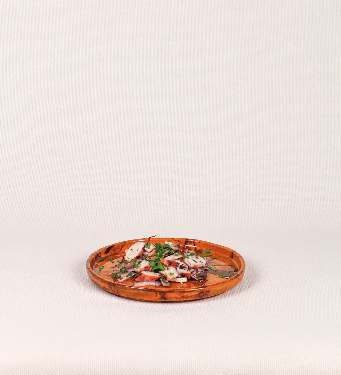 Prato de aperitivos com design minimalista e distinto. O prato é de barro, com uma mistura de dois tipos de barro diferentes, um mais escuro e outro mais claro. Esta mistura cria um desenho de formas abstratas e aleatórias, que se espalham por todas as superfícies do prato. Tem acabamento vidrado castanho translúcido, escurecendo as cores originais dos barros misturados. A forma do prato é circular, com uma borda envolvente de altura baixa e design simples e minimalista. 