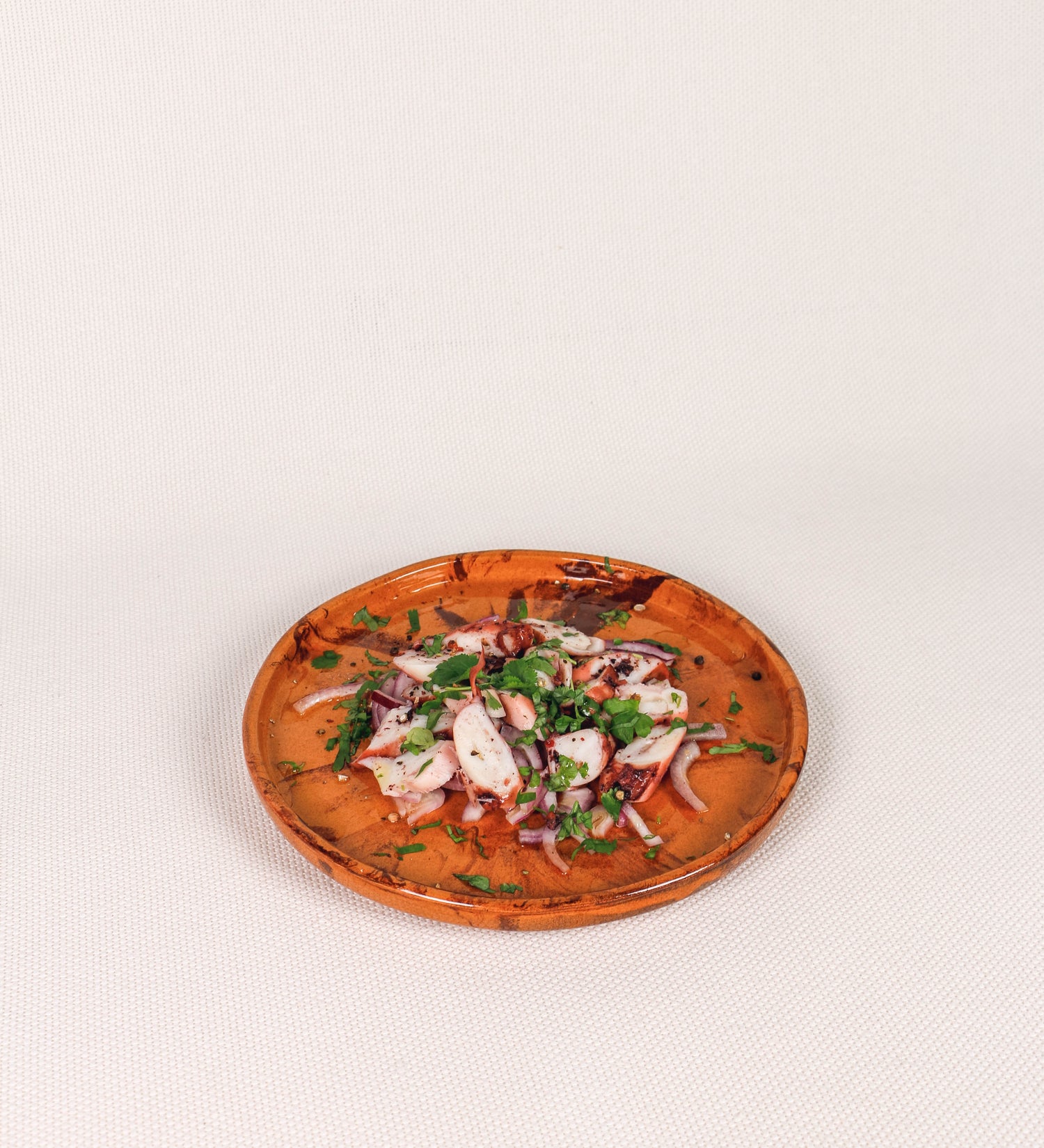 Prato de aperitivos com design minimalista e distinto. O prato é de barro, com uma mistura de dois tipos de barro diferentes, um mais escuro e outro mais claro. Esta mistura cria um desenho de formas abstratas e aleatórias, que se espalham por todas as superfícies do prato. Tem acabamento vidrado castanho translúcido, escurecendo as cores originais dos barros misturados. A forma do prato é circular, com uma borda envolvente de altura baixa e design simples e minimalista. 