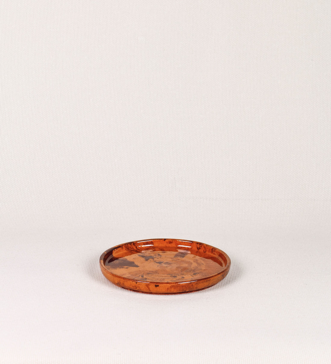 Prato de sobremesa com design minimalista e distinto. O prato é de barro, com uma mistura de dois tipos de barro diferentes, um mais escuro e outro mais claro. Esta mistura cria um desenho de formas abstratas e aleatórias, que se espalham por todas as superfícies do prato. Tem acabamento vidrado castanho translúcido, escurecendo as cores originais dos barros misturados. A forma do prato é circular, com uma borda envolvente de altura baixa e design simples e minimalista. 