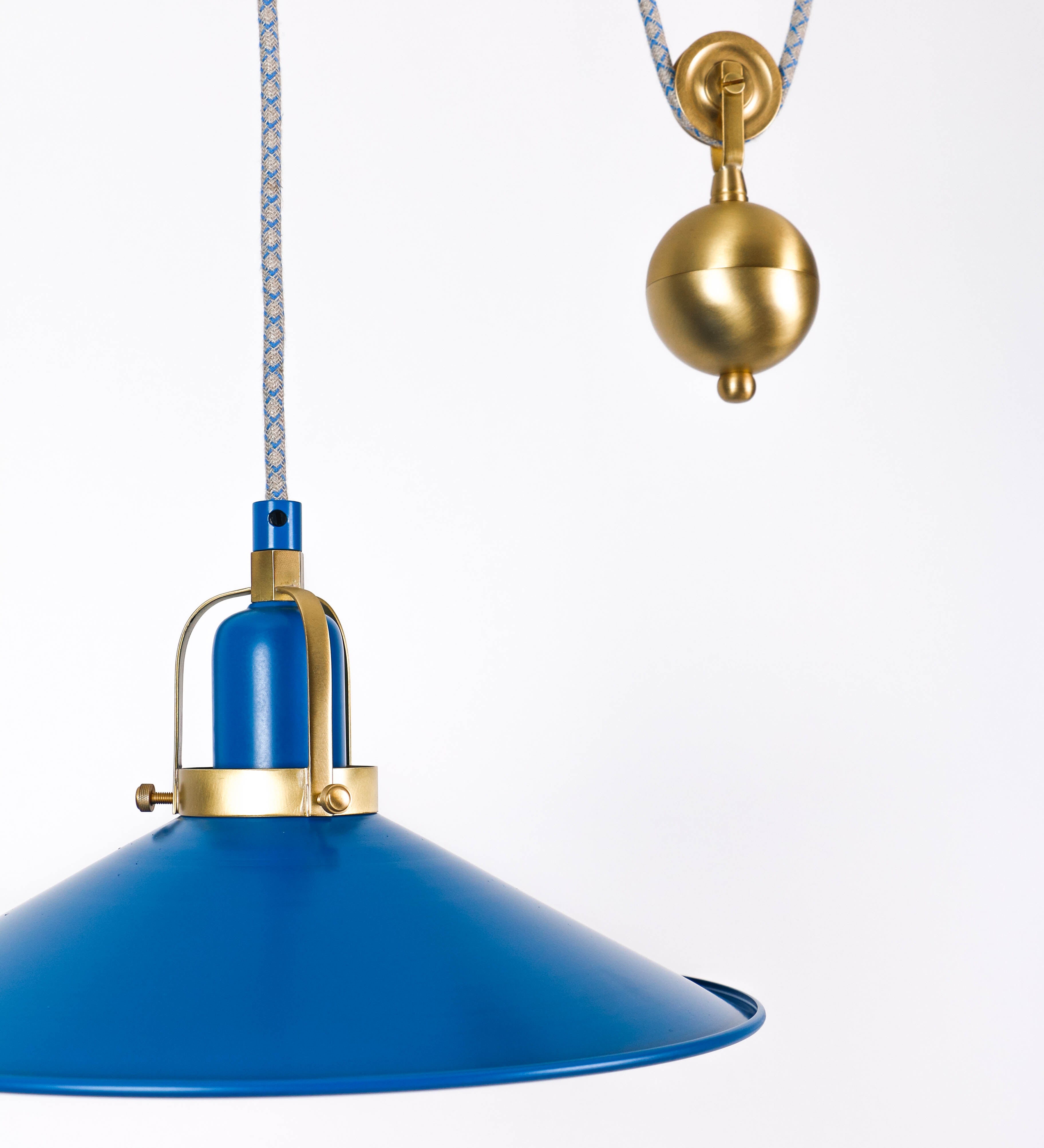 Suspensão de teto composta por uma base circular azul vintage de onde desce um cabo com padrão de linhas azuis e beges. O cabo passa por uma roldana em latão escovado fixa ao teto, e desce até ao chapéu. Entre a base e a roldana o cabo sustenta uma esfera em latão escovado, que atua como um contrapeso, para ajustar a altura. O chapéu, azul vintage, tem formato cónico invertido, uma borda ligeiramente levantada e interior branco. No topo do chapéu há um suporte em latão escovado que conecta o chapéu ao cabo.
