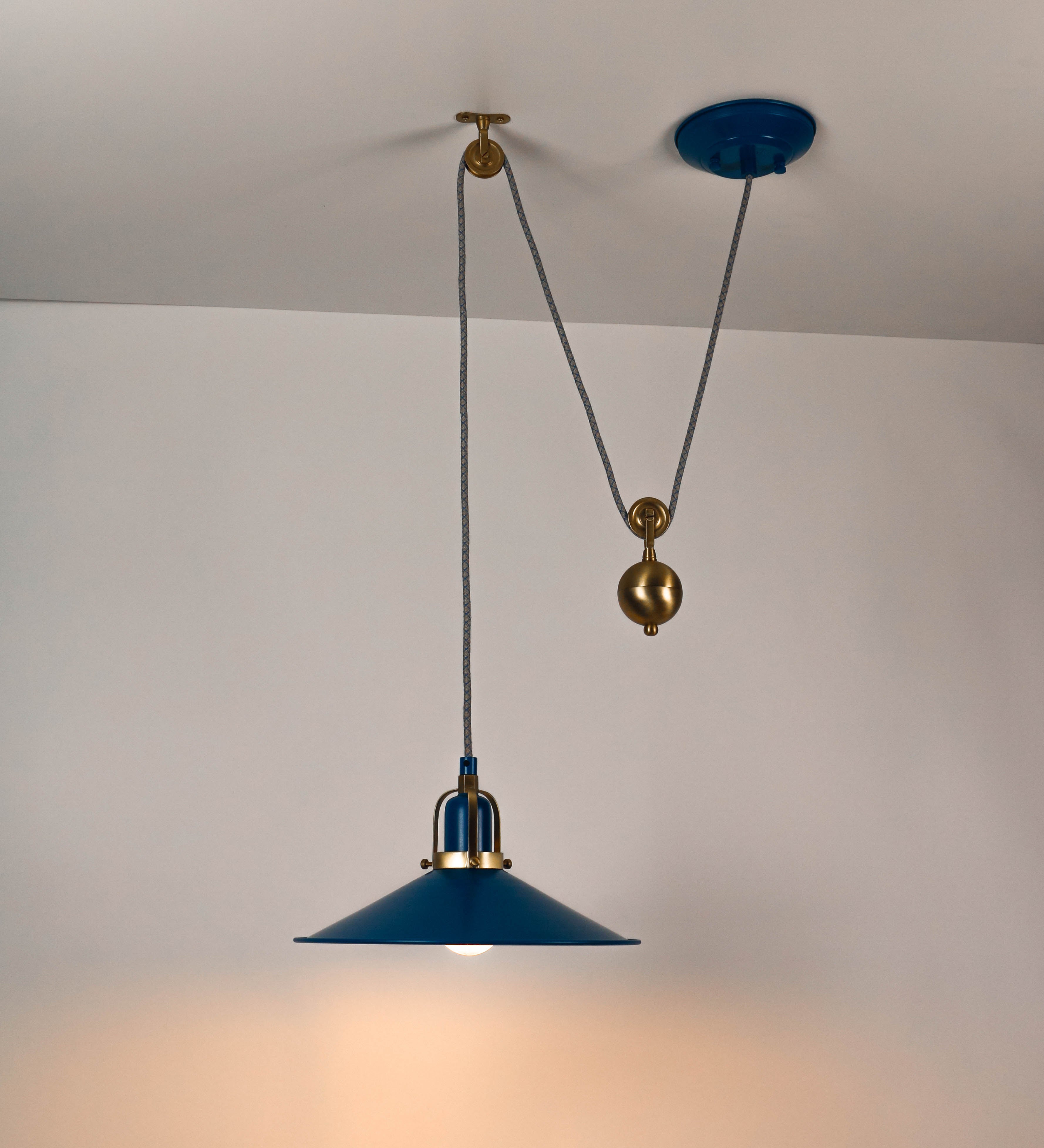 Suspensão de teto composta por uma base circular azul vintage de onde desce um cabo com padrão de linhas azuis e beges. O cabo passa por uma roldana em latão escovado fixa ao teto, e desce até ao chapéu. Entre a base e a roldana o cabo sustenta uma esfera em latão escovado, que atua como um contrapeso, para ajustar a altura. O chapéu, azul vintage, tem formato cónico invertido, uma borda ligeiramente levantada e interior branco. No topo do chapéu há um suporte em latão escovado que conecta o chapéu ao cabo.