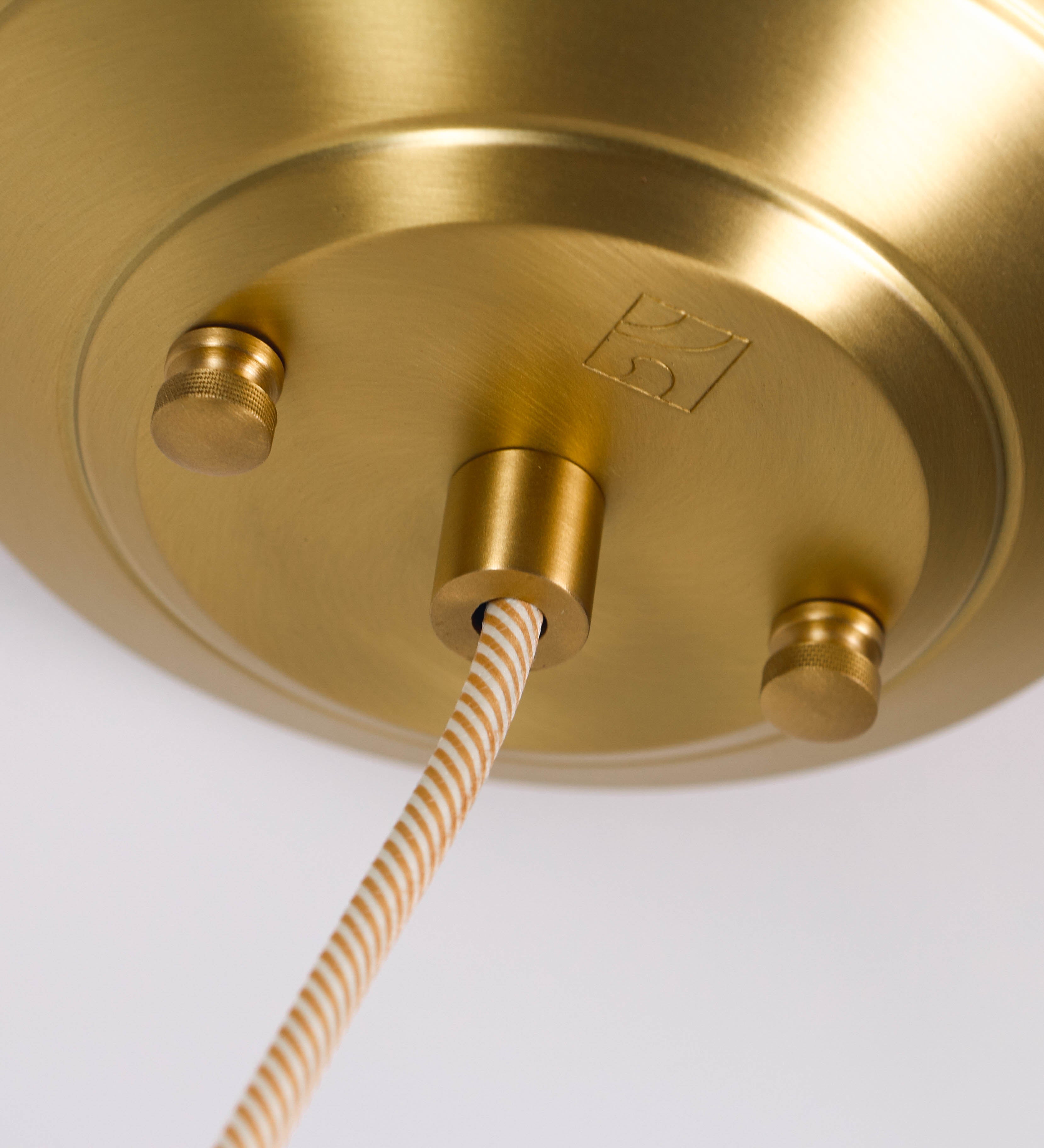 Suspensão de teto em latão escovado, composta por uma base circular, de onde desce um cabo revestido têxtil com um padrão com linhas douradas e brancas. O cabo passa por uma roldana fixa ao teto, e desce até ao chapéu. Entre a base e a roldana, o cabo sustenta uma esfera, que atua como um contrapeso, para ajustar a altura do candeeiro. O chapéu tem formato cónico invertido e uma borda ligeiramente levantada. No topo do chapéu, há um suporte circular com três pequenos braços que conecta o chapéu ao cabo.