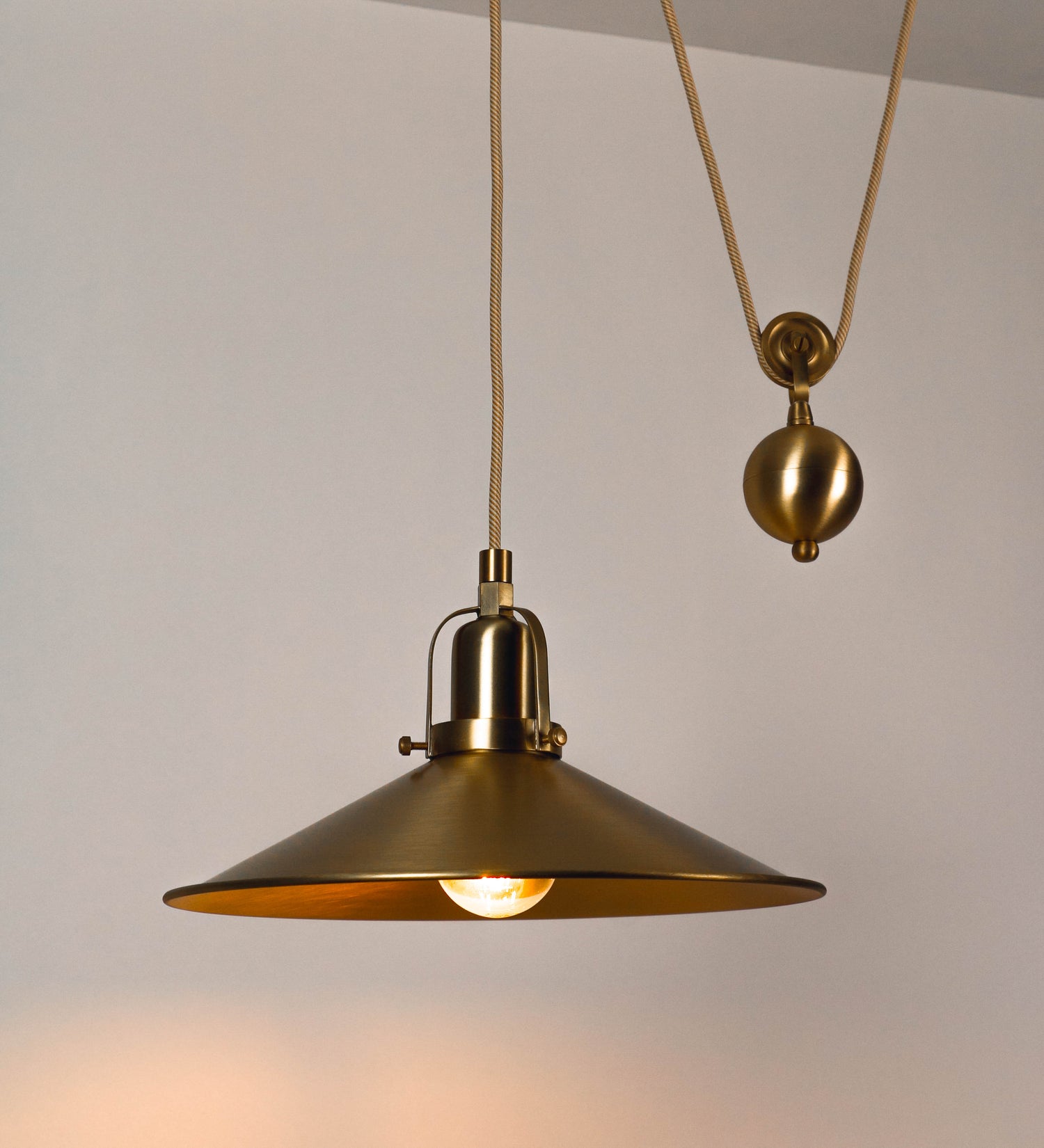 Suspensão de teto em latão escovado, composta por uma base circular, de onde desce um cabo revestido têxtil com um padrão com linhas douradas e brancas. O cabo passa por uma roldana fixa ao teto, e desce até ao chapéu. Entre a base e a roldana, o cabo sustenta uma esfera, que atua como um contrapeso, para ajustar a altura do candeeiro. O chapéu tem formato cónico invertido e uma borda ligeiramente levantada. No topo do chapéu, há um suporte circular com três pequenos braços que conecta o chapéu ao cabo.