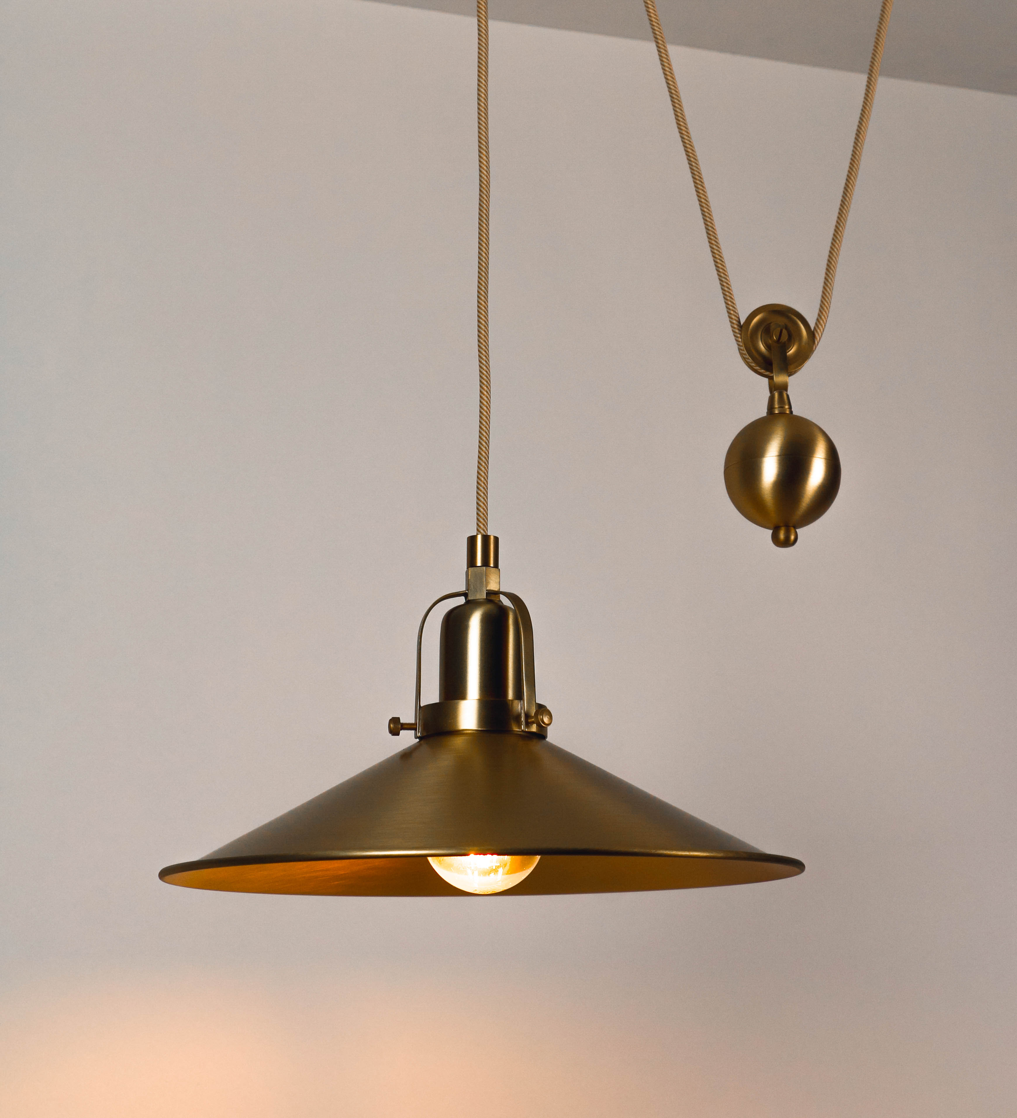 Suspensão de teto em latão escovado, composta por uma base circular, de onde desce um cabo revestido têxtil com um padrão com linhas douradas e brancas. O cabo passa por uma roldana fixa ao teto, e desce até ao chapéu. Entre a base e a roldana, o cabo sustenta uma esfera, que atua como um contrapeso, para ajustar a altura do candeeiro. O chapéu tem formato cónico invertido e uma borda ligeiramente levantada. No topo do chapéu, há um suporte circular com três pequenos braços que conecta o chapéu ao cabo.