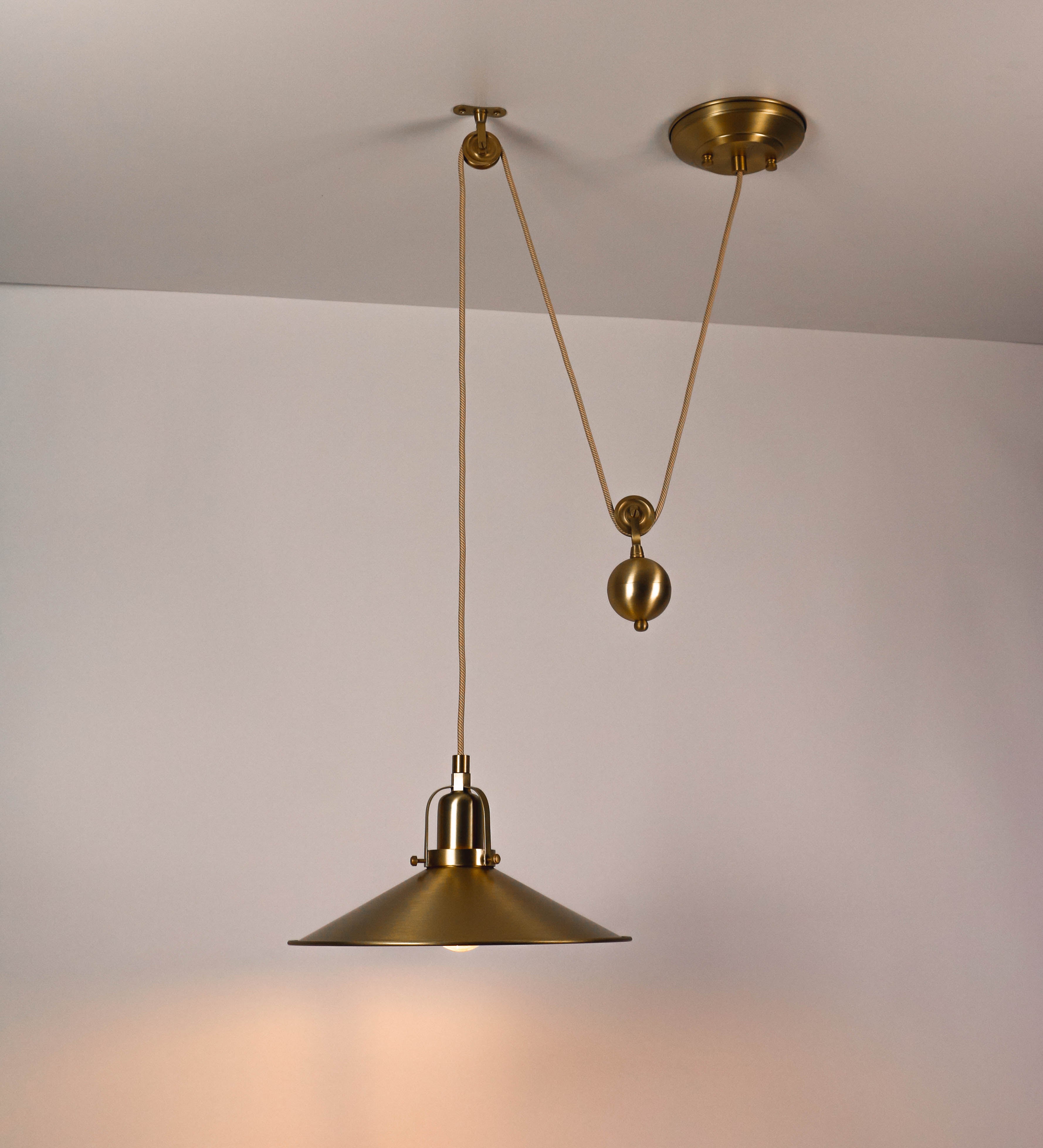 Suspensão de teto em latão escovado, composta por uma base circular, de onde desce um cabo revestido têxtil com um padrão com linhas douradas e brancas. O cabo passa por uma roldana fixa ao teto, e desce até ao chapéu. Entre a base e a roldana, o cabo sustenta uma esfera, que atua como um contrapeso, para ajustar a altura do candeeiro. O chapéu tem formato cónico invertido e uma borda ligeiramente levantada. No topo do chapéu, há um suporte circular com três pequenos braços que conecta o chapéu ao cabo.