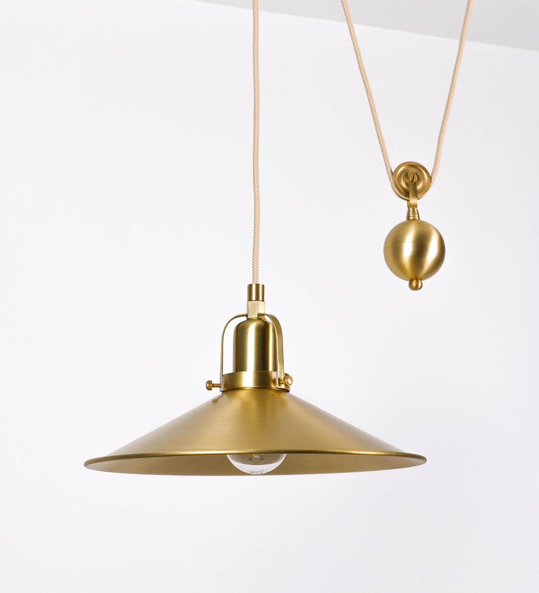 Suspensão de teto em latão escovado, composta por uma base circular, de onde desce um cabo revestido têxtil com um padrão com linhas douradas e brancas. O cabo passa por uma roldana fixa ao teto, e desce até ao chapéu. Entre a base e a roldana, o cabo sustenta uma esfera, que atua como um contrapeso, para ajustar a altura do candeeiro. O chapéu tem formato cónico invertido e uma borda ligeiramente levantada. No topo do chapéu, há um suporte circular com três pequenos braços que conecta o chapéu ao cabo.