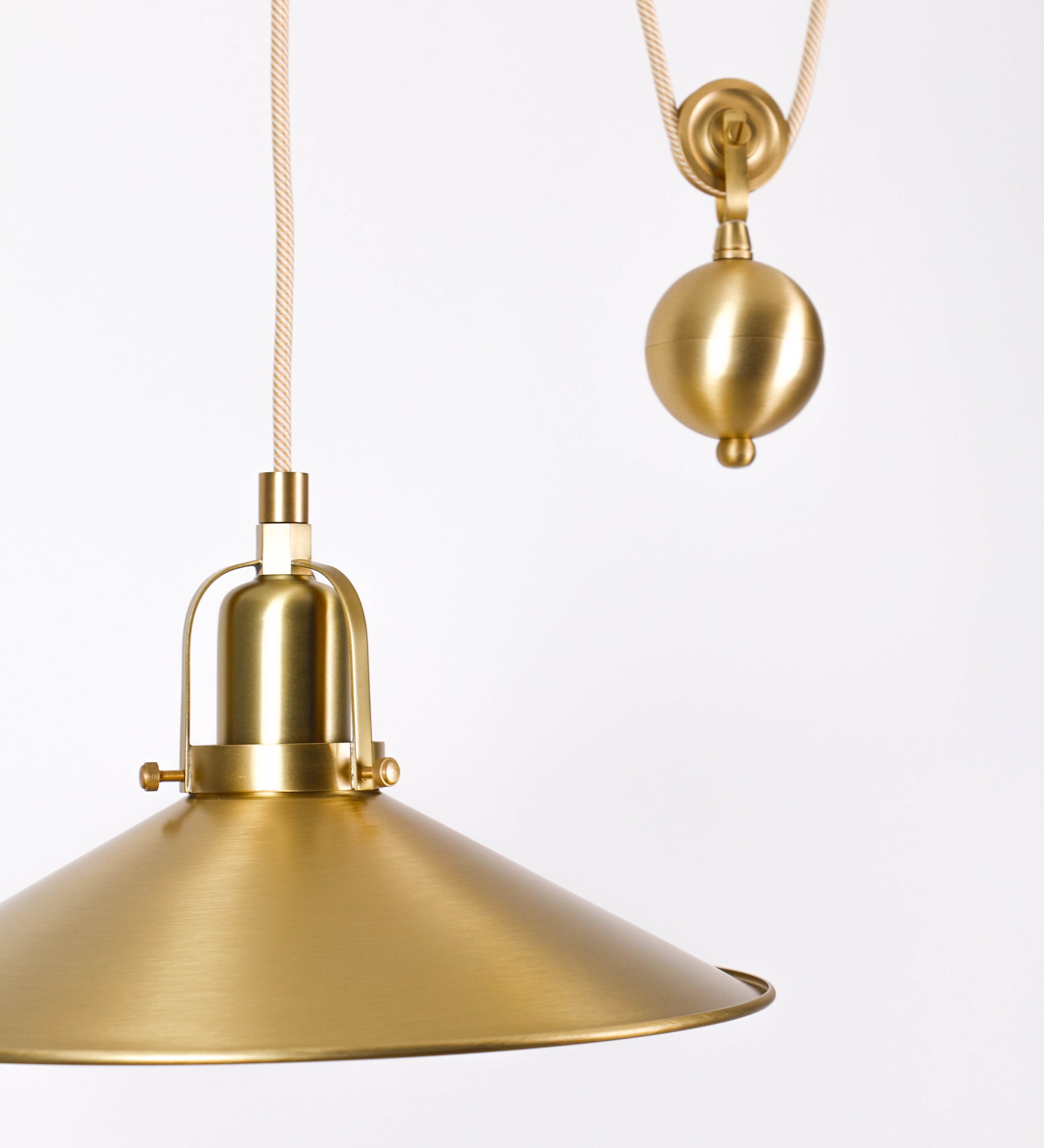 Suspensão de teto em latão escovado, composta por uma base circular, de onde desce um cabo revestido têxtil com um padrão com linhas douradas e brancas. O cabo passa por uma roldana fixa ao teto, e desce até ao chapéu. Entre a base e a roldana, o cabo sustenta uma esfera, que atua como um contrapeso, para ajustar a altura do candeeiro. O chapéu tem formato cónico invertido e uma borda ligeiramente levantada. No topo do chapéu, há um suporte circular com três pequenos braços que conecta o chapéu ao cabo.