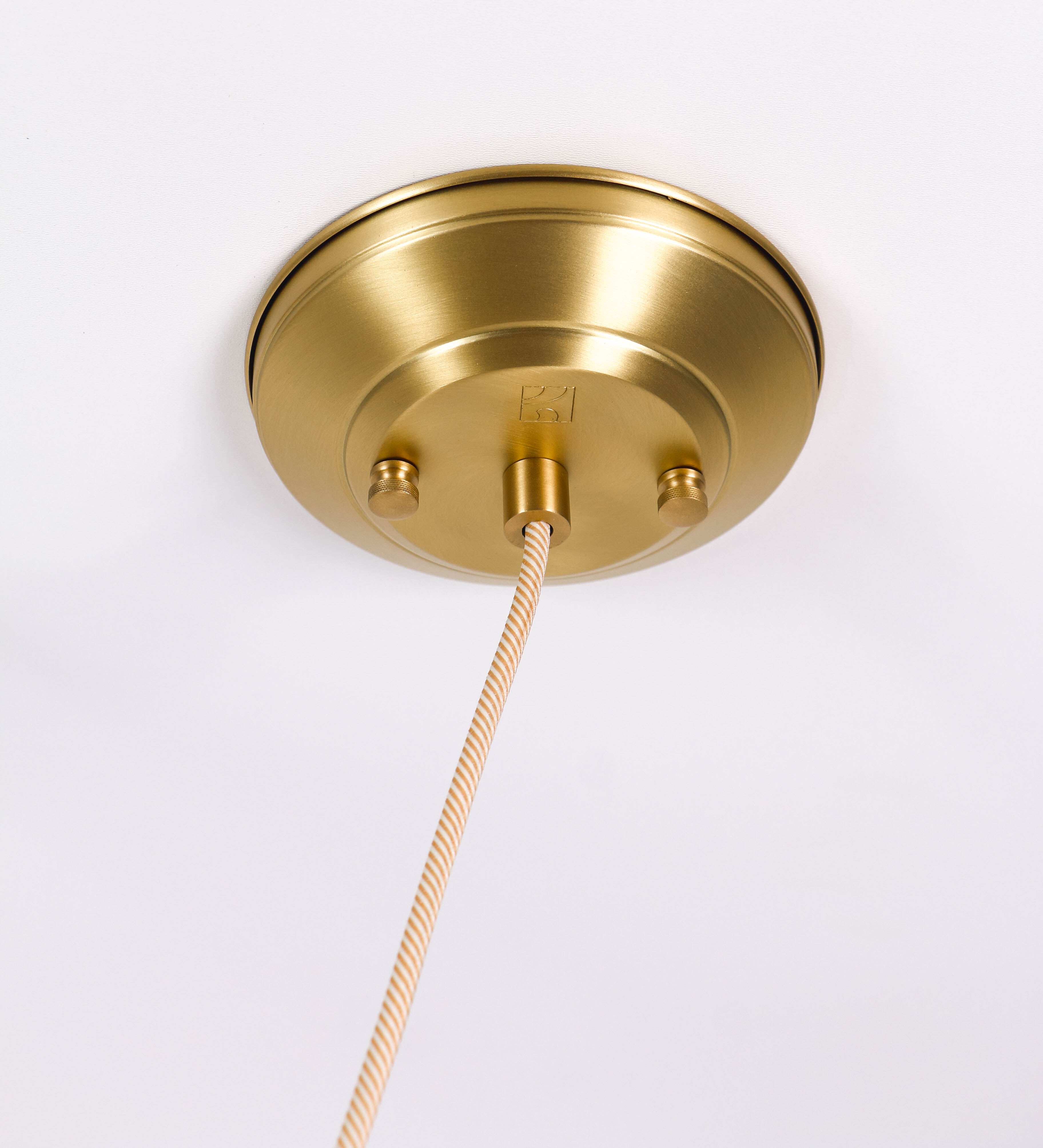 Suspensão de teto em latão escovado, composta por uma base circular, de onde desce um cabo revestido têxtil com um padrão com linhas douradas e brancas. O cabo passa por uma roldana fixa ao teto, e desce até ao chapéu. Entre a base e a roldana, o cabo sustenta uma esfera, que atua como um contrapeso, para ajustar a altura do candeeiro. O chapéu tem formato cónico invertido e uma borda ligeiramente levantada. No topo do chapéu, há um suporte circular com três pequenos braços que conecta o chapéu ao cabo.