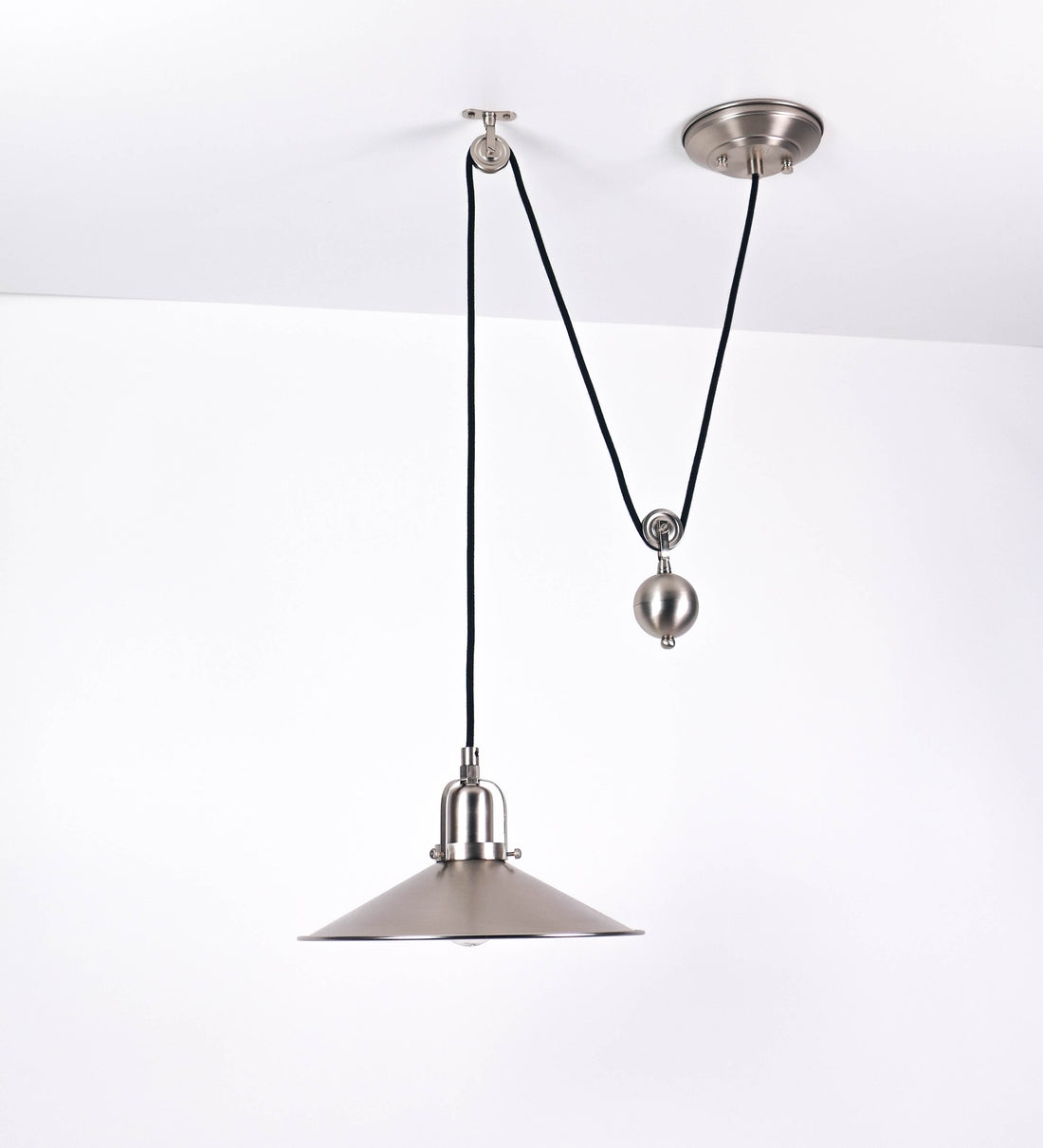 Suspensão de teto em níquel escovado, composta por uma base circular, de onde desce um cabo revestido têxtil preto. O cabo passa por uma roldana fixa ao teto, e desce até ao chapéu. Entre a base e a roldana, o cabo sustenta uma esfera, que atua como um contrapeso, para ajustar a altura do candeeiro. O chapéu tem formato cónico invertido e uma borda ligeiramente levantada. No topo do chapéu, há um suporte circular com três pequenos braços que conecta o chapéu ao cabo.