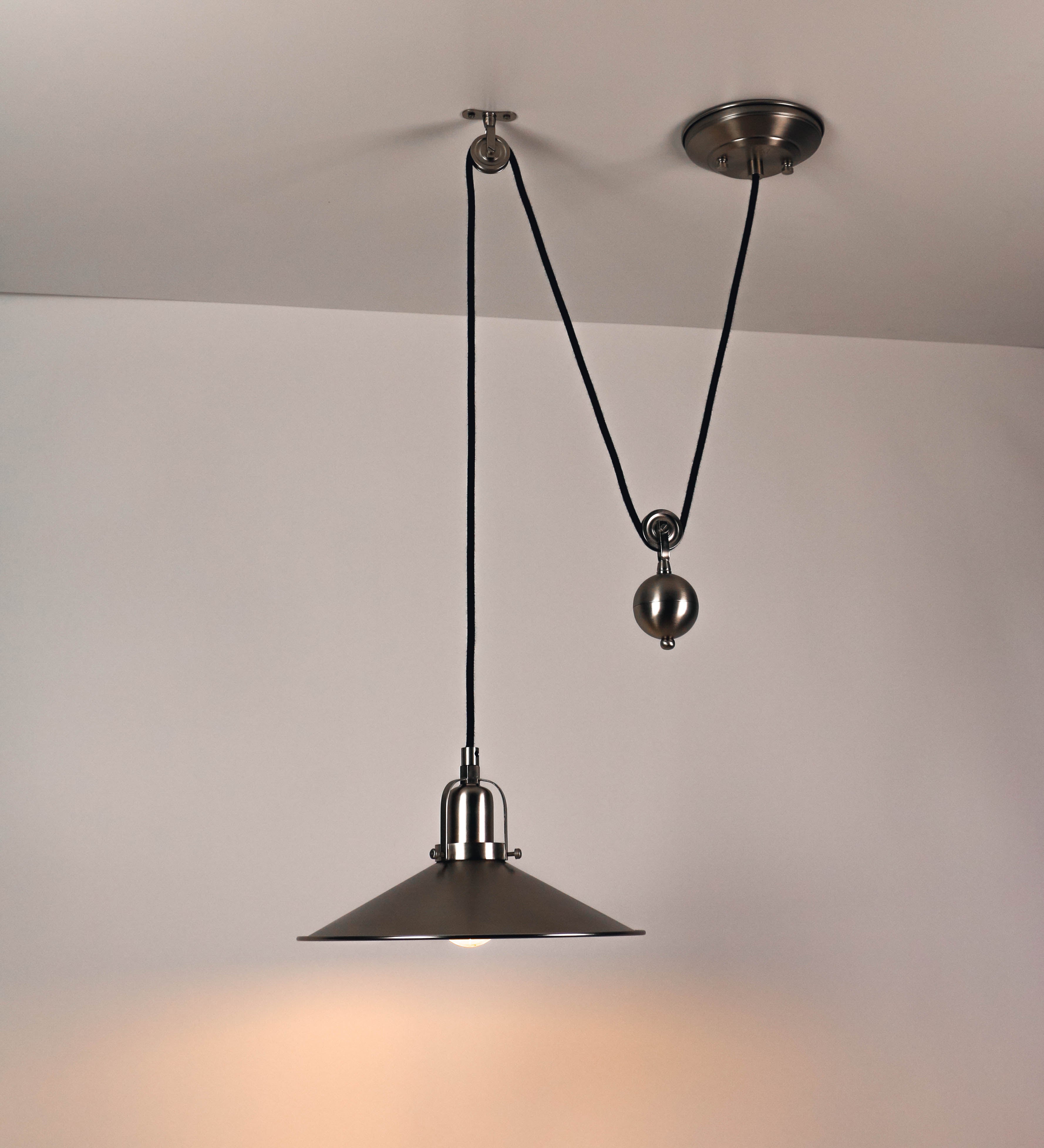 Suspensão de teto em níquel escovado, composta por uma base circular, de onde desce um cabo revestido têxtil preto. O cabo passa por uma roldana fixa ao teto, e desce até ao chapéu. Entre a base e a roldana, o cabo sustenta uma esfera, que atua como um contrapeso, para ajustar a altura do candeeiro. O chapéu tem formato cónico invertido e uma borda ligeiramente levantada. No topo do chapéu, há um suporte circular com três pequenos braços que conecta o chapéu ao cabo.
