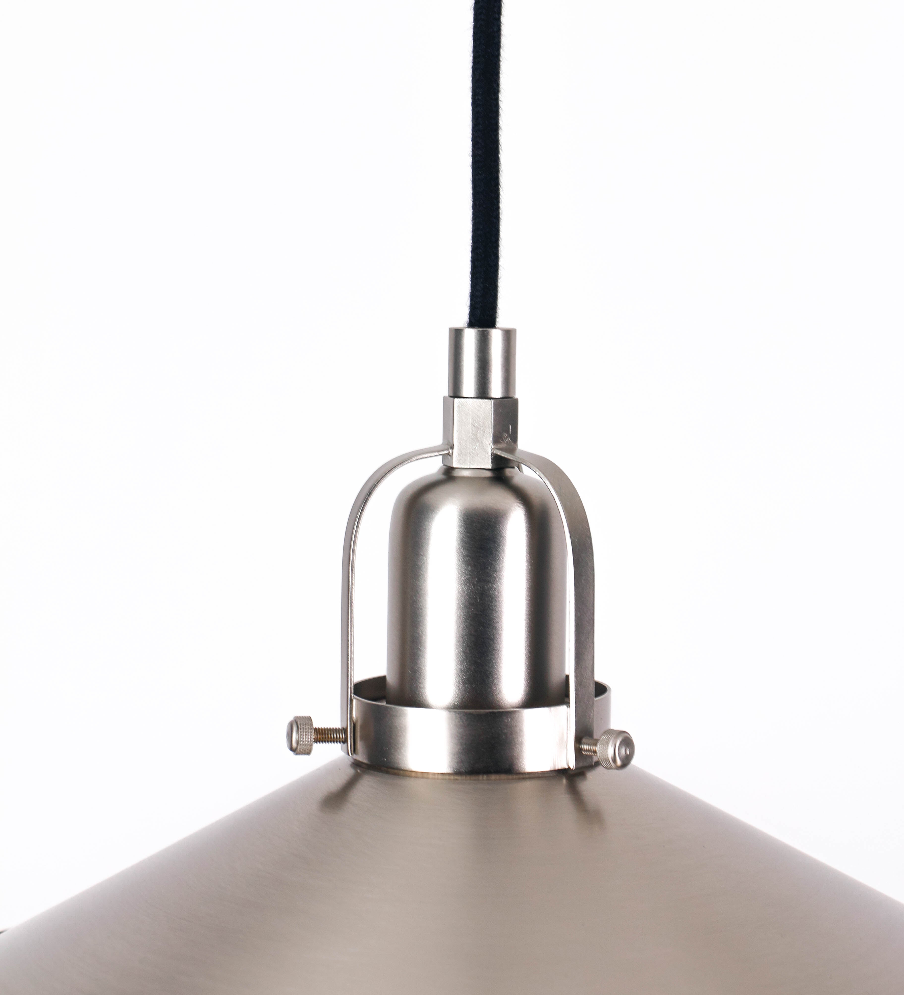 Suspensão de teto em níquel escovado, composta por uma base circular, de onde desce um cabo revestido têxtil preto. O cabo passa por uma roldana fixa ao teto, e desce até ao chapéu. Entre a base e a roldana, o cabo sustenta uma esfera, que atua como um contrapeso, para ajustar a altura do candeeiro. O chapéu tem formato cónico invertido e uma borda ligeiramente levantada. No topo do chapéu, há um suporte circular com três pequenos braços que conecta o chapéu ao cabo.