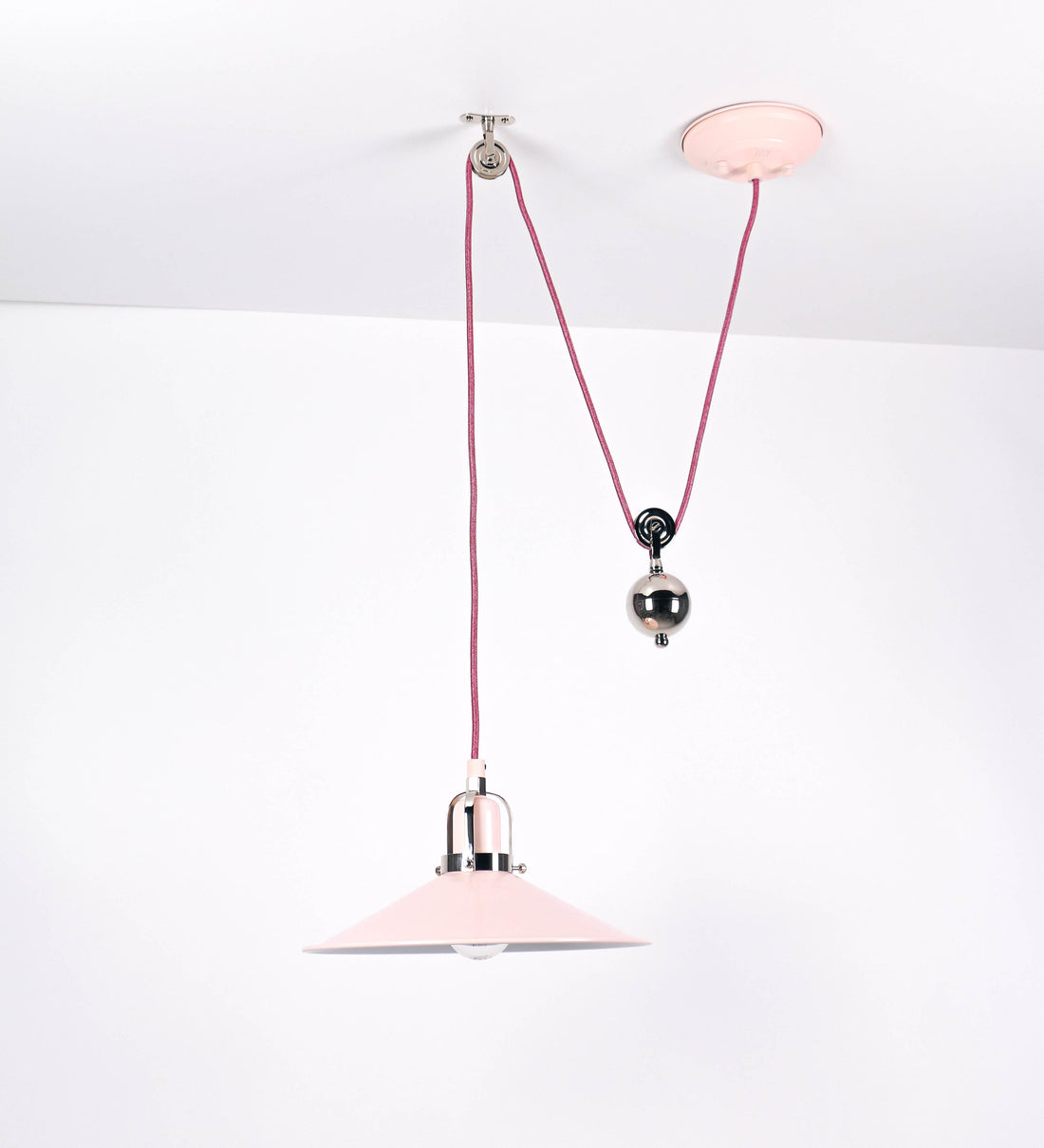 Suspensão de teto composta por uma base circular rosa clara, de onde desce um cabo revestido têxtil rosa velho. O cabo passa por uma roldana em níquel polido, fixa ao teto, e desce até ao chapéu. Entre a base e a roldana, o cabo sustenta uma esfera, em níquel polido, que atua como um contrapeso, para ajustar a altura. O chapéu, rosa claro, tem formato cónico invertido, uma borda ligeiramente levantada e interior branco. No topo do chapéu, há um suporte em níquel polido que conecta o chapéu ao cabo.