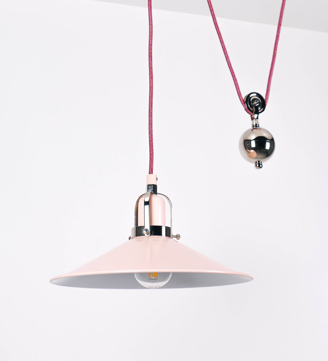 Suspensão de teto composta por uma base circular rosa clara, de onde desce um cabo revestido têxtil rosa velho. O cabo passa por uma roldana em níquel polido, fixa ao teto, e desce até ao chapéu. Entre a base e a roldana, o cabo sustenta uma esfera, em níquel polido, que atua como um contrapeso, para ajustar a altura. O chapéu, rosa claro, tem formato cónico invertido, uma borda ligeiramente levantada e interior branco. No topo do chapéu, há um suporte em níquel polido que conecta o chapéu ao cabo.