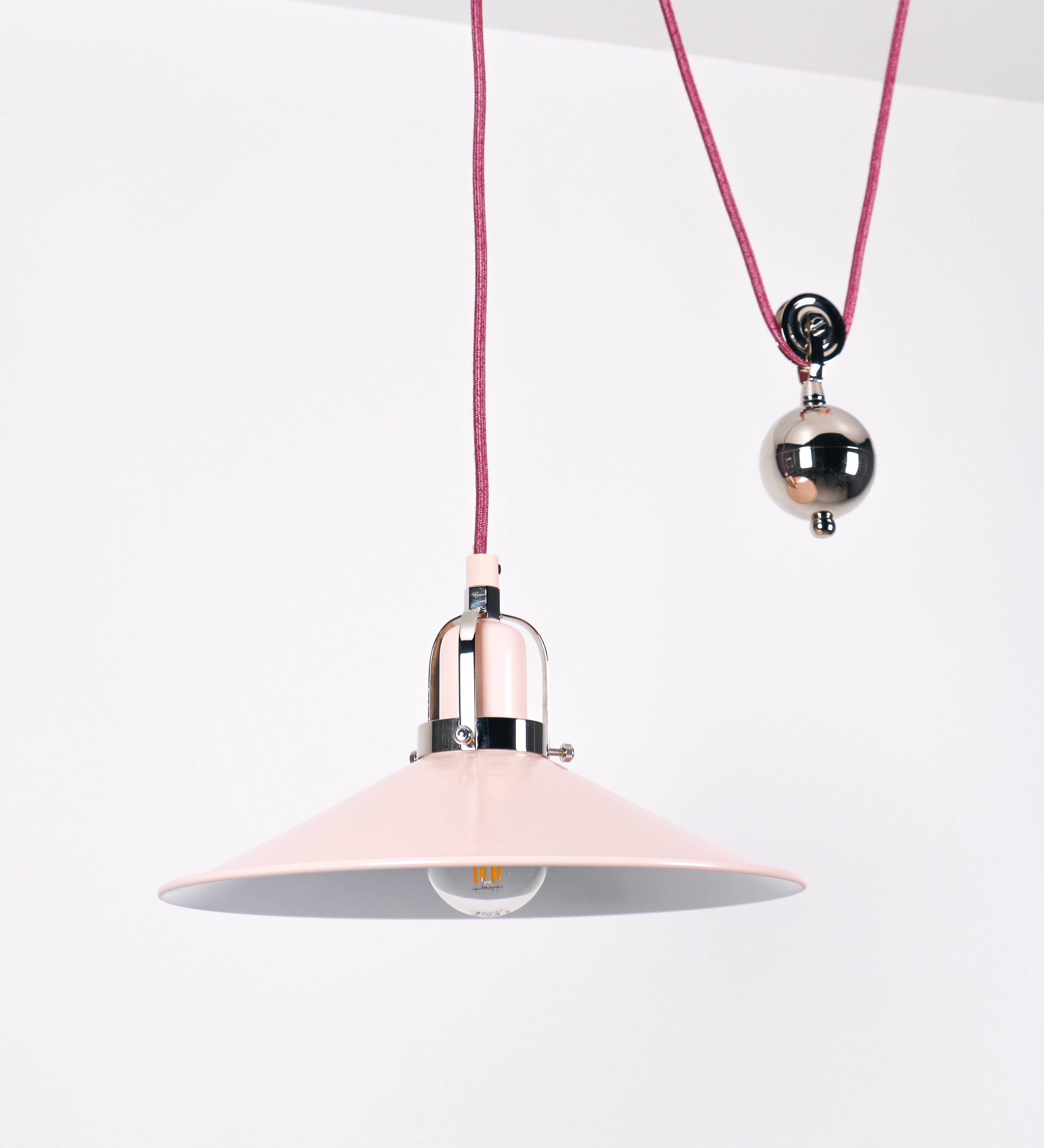 Suspensão de teto composta por uma base circular rosa clara, de onde desce um cabo revestido têxtil rosa velho. O cabo passa por uma roldana em níquel polido, fixa ao teto, e desce até ao chapéu. Entre a base e a roldana, o cabo sustenta uma esfera, em níquel polido, que atua como um contrapeso, para ajustar a altura. O chapéu, rosa claro, tem formato cónico invertido, uma borda ligeiramente levantada e interior branco. No topo do chapéu, há um suporte em níquel polido que conecta o chapéu ao cabo.