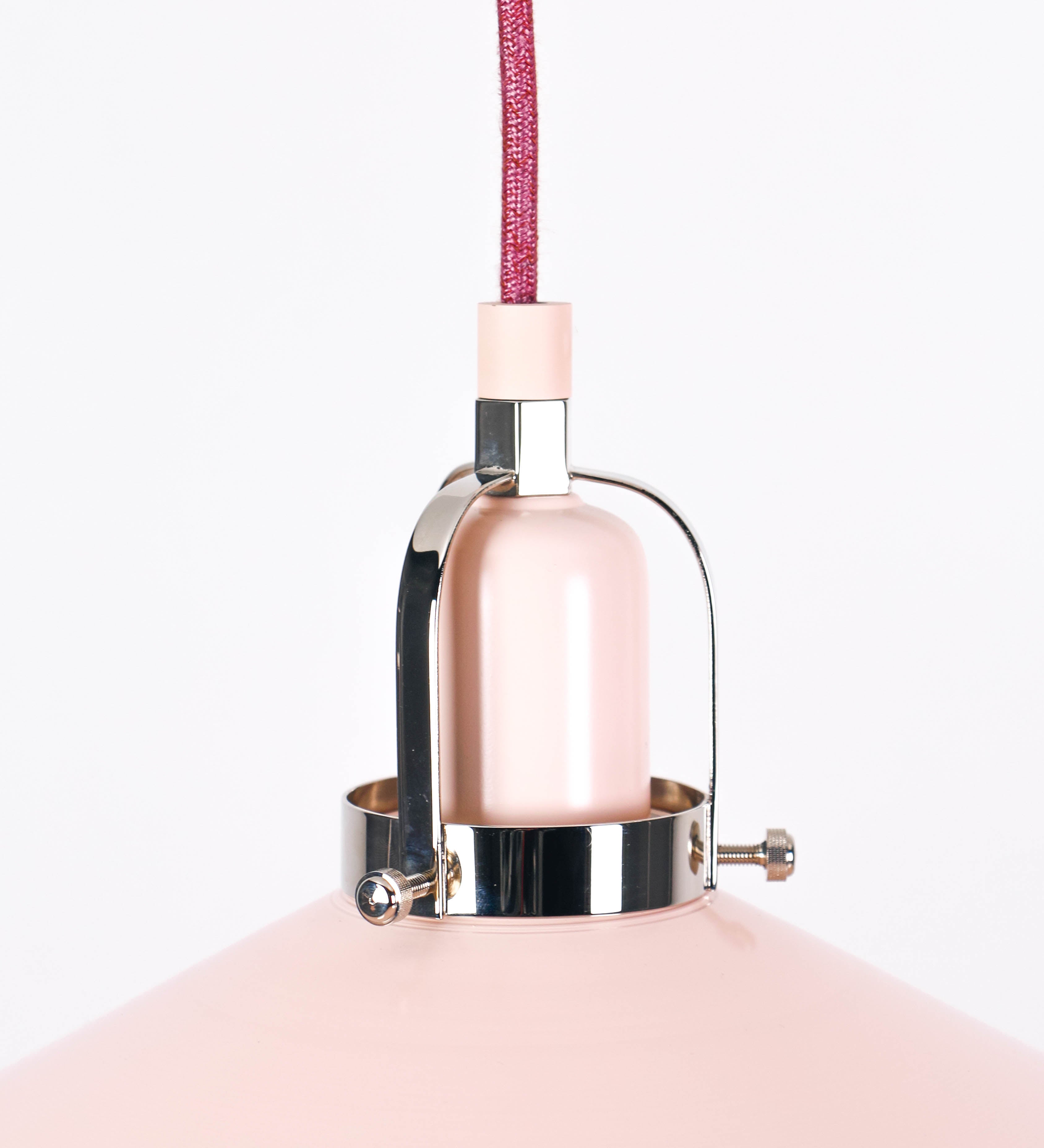 Suspensão de teto composta por uma base circular rosa clara, de onde desce um cabo revestido têxtil rosa velho. O cabo passa por uma roldana em níquel polido, fixa ao teto, e desce até ao chapéu. Entre a base e a roldana, o cabo sustenta uma esfera, em níquel polido, que atua como um contrapeso, para ajustar a altura. O chapéu, rosa claro, tem formato cónico invertido, uma borda ligeiramente levantada e interior branco. No topo do chapéu, há um suporte em níquel polido que conecta o chapéu ao cabo.