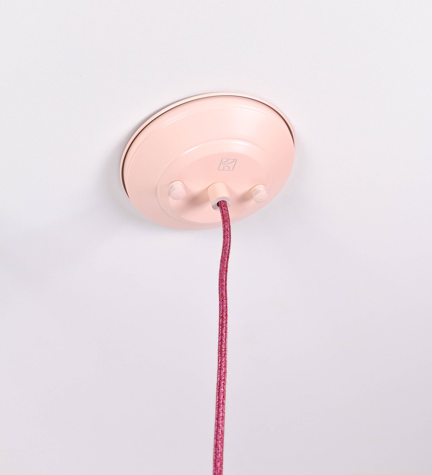 Suspensão de teto composta por uma base circular rosa clara, de onde desce um cabo revestido têxtil rosa velho. O cabo passa por uma roldana em níquel polido, fixa ao teto, e desce até ao chapéu. Entre a base e a roldana, o cabo sustenta uma esfera, em níquel polido, que atua como um contrapeso, para ajustar a altura. O chapéu, rosa claro, tem formato cónico invertido, uma borda ligeiramente levantada e interior branco. No topo do chapéu, há um suporte em níquel polido que conecta o chapéu ao cabo.
