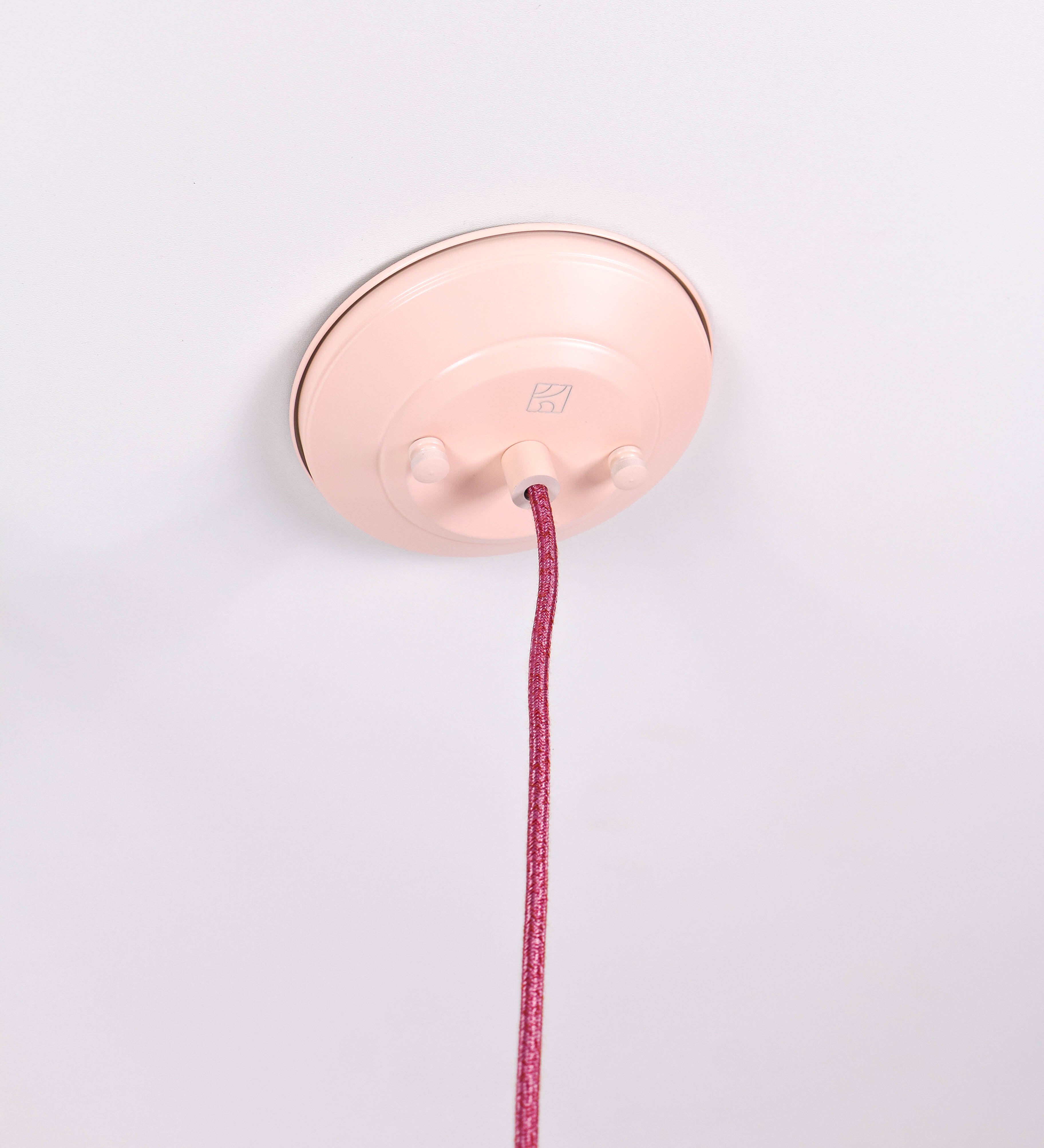 Suspensão de teto composta por uma base circular rosa clara, de onde desce um cabo revestido têxtil rosa velho. O cabo passa por uma roldana em níquel polido, fixa ao teto, e desce até ao chapéu. Entre a base e a roldana, o cabo sustenta uma esfera, em níquel polido, que atua como um contrapeso, para ajustar a altura. O chapéu, rosa claro, tem formato cónico invertido, uma borda ligeiramente levantada e interior branco. No topo do chapéu, há um suporte em níquel polido que conecta o chapéu ao cabo.