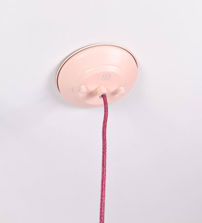 Suspensão de teto composta por uma base circular rosa clara, de onde desce um cabo revestido têxtil rosa velho. O cabo passa por uma roldana em níquel polido, fixa ao teto, e desce até ao chapéu. Entre a base e a roldana, o cabo sustenta uma esfera, em níquel polido, que atua como um contrapeso, para ajustar a altura. O chapéu, rosa claro, tem formato cónico invertido, uma borda ligeiramente levantada e interior branco. No topo do chapéu, há um suporte em níquel polido que conecta o chapéu ao cabo.