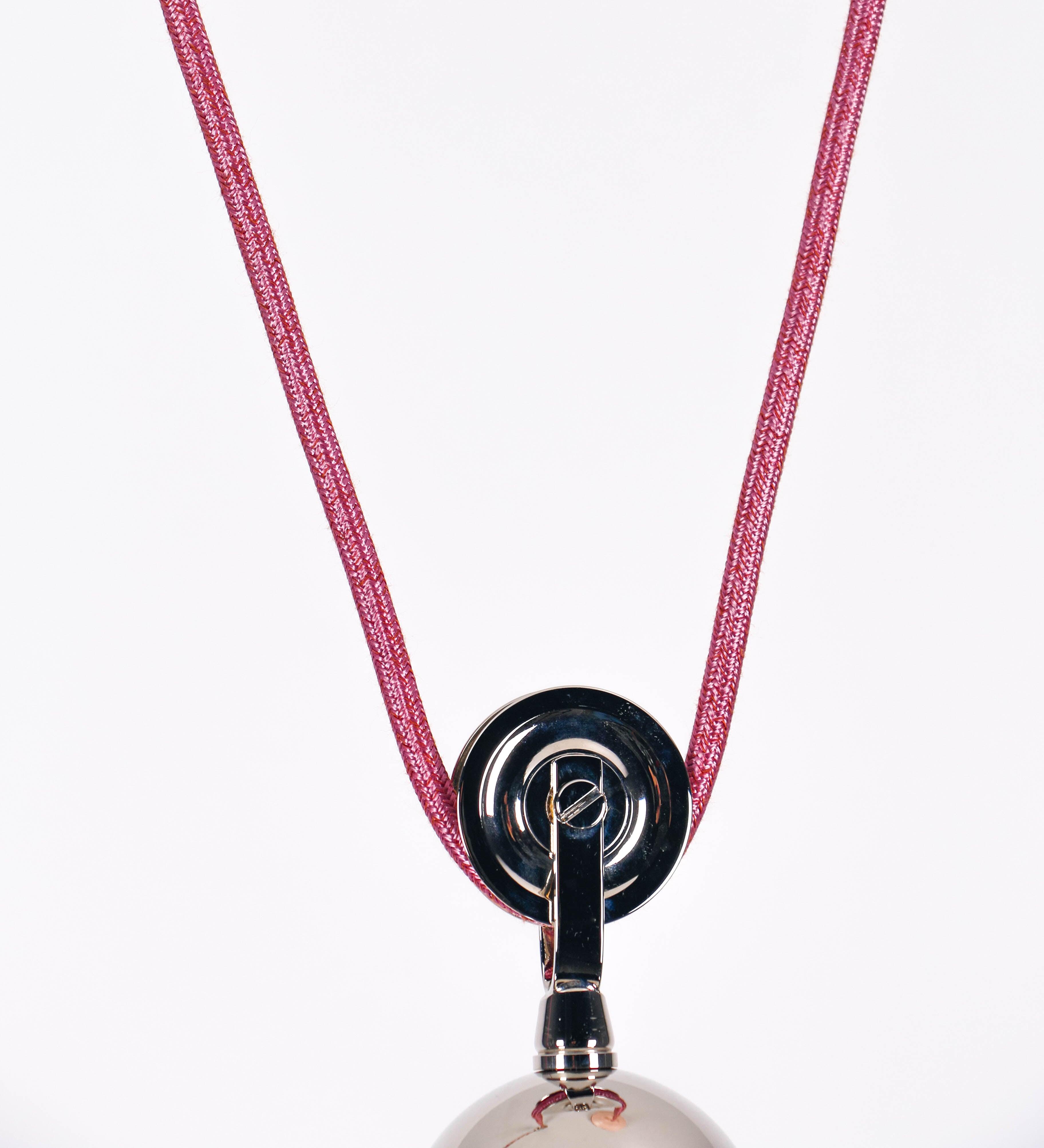 Suspensão de teto composta por uma base circular rosa clara, de onde desce um cabo revestido têxtil rosa velho. O cabo passa por uma roldana em níquel polido, fixa ao teto, e desce até ao chapéu. Entre a base e a roldana, o cabo sustenta uma esfera, em níquel polido, que atua como um contrapeso, para ajustar a altura. O chapéu, rosa claro, tem formato cónico invertido, uma borda ligeiramente levantada e interior branco. No topo do chapéu, há um suporte em níquel polido que conecta o chapéu ao cabo.