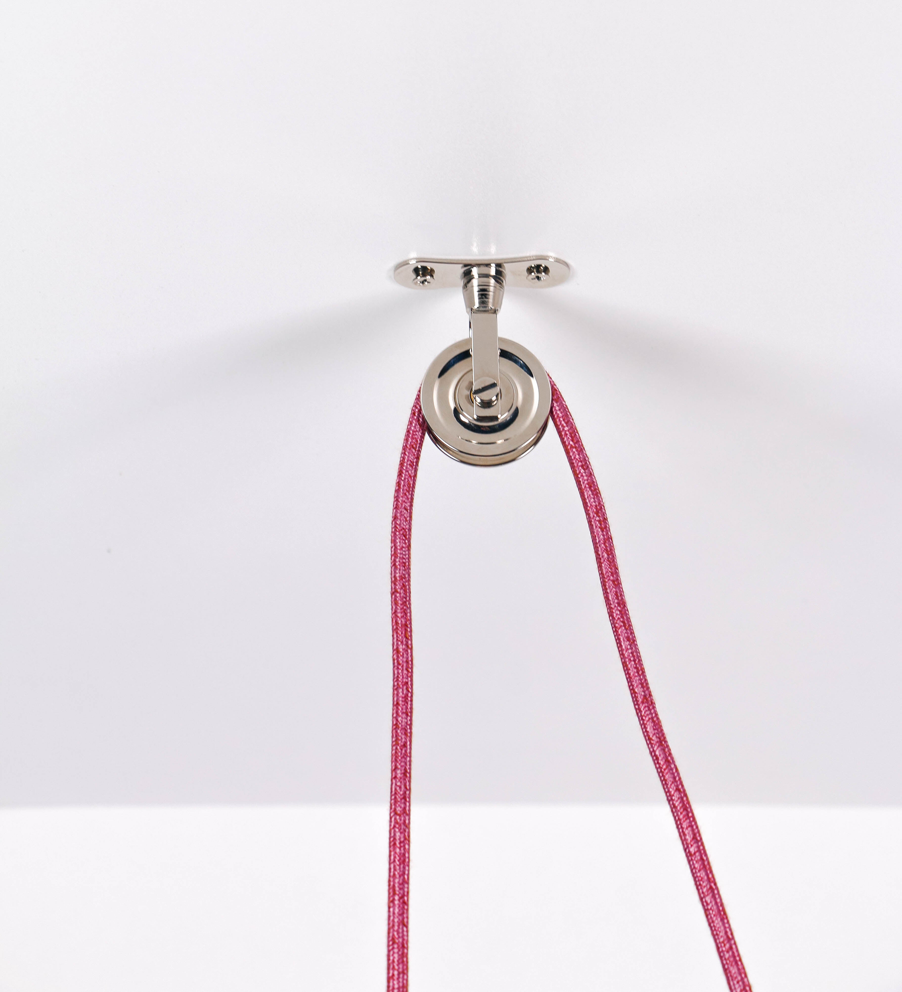 Suspensão de teto composta por uma base circular rosa clara, de onde desce um cabo revestido têxtil rosa velho. O cabo passa por uma roldana em níquel polido, fixa ao teto, e desce até ao chapéu. Entre a base e a roldana, o cabo sustenta uma esfera, em níquel polido, que atua como um contrapeso, para ajustar a altura. O chapéu, rosa claro, tem formato cónico invertido, uma borda ligeiramente levantada e interior branco. No topo do chapéu, há um suporte em níquel polido que conecta o chapéu ao cabo.