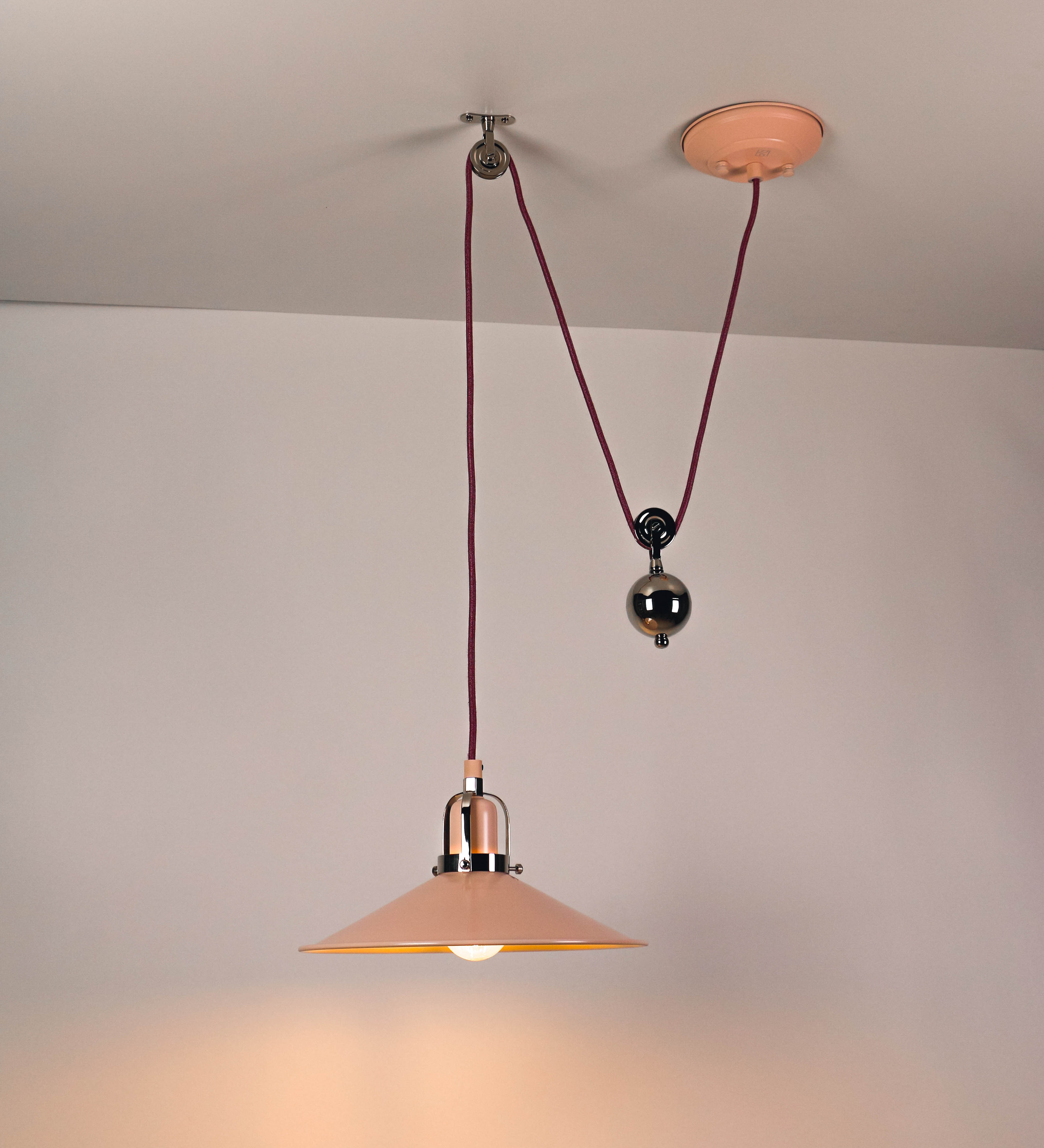 Suspensão de teto composta por uma base circular rosa clara, de onde desce um cabo revestido têxtil rosa velho. O cabo passa por uma roldana em níquel polido, fixa ao teto, e desce até ao chapéu. Entre a base e a roldana, o cabo sustenta uma esfera, em níquel polido, que atua como um contrapeso, para ajustar a altura. O chapéu, rosa claro, tem formato cónico invertido, uma borda ligeiramente levantada e interior branco. No topo do chapéu, há um suporte em níquel polido que conecta o chapéu ao cabo.