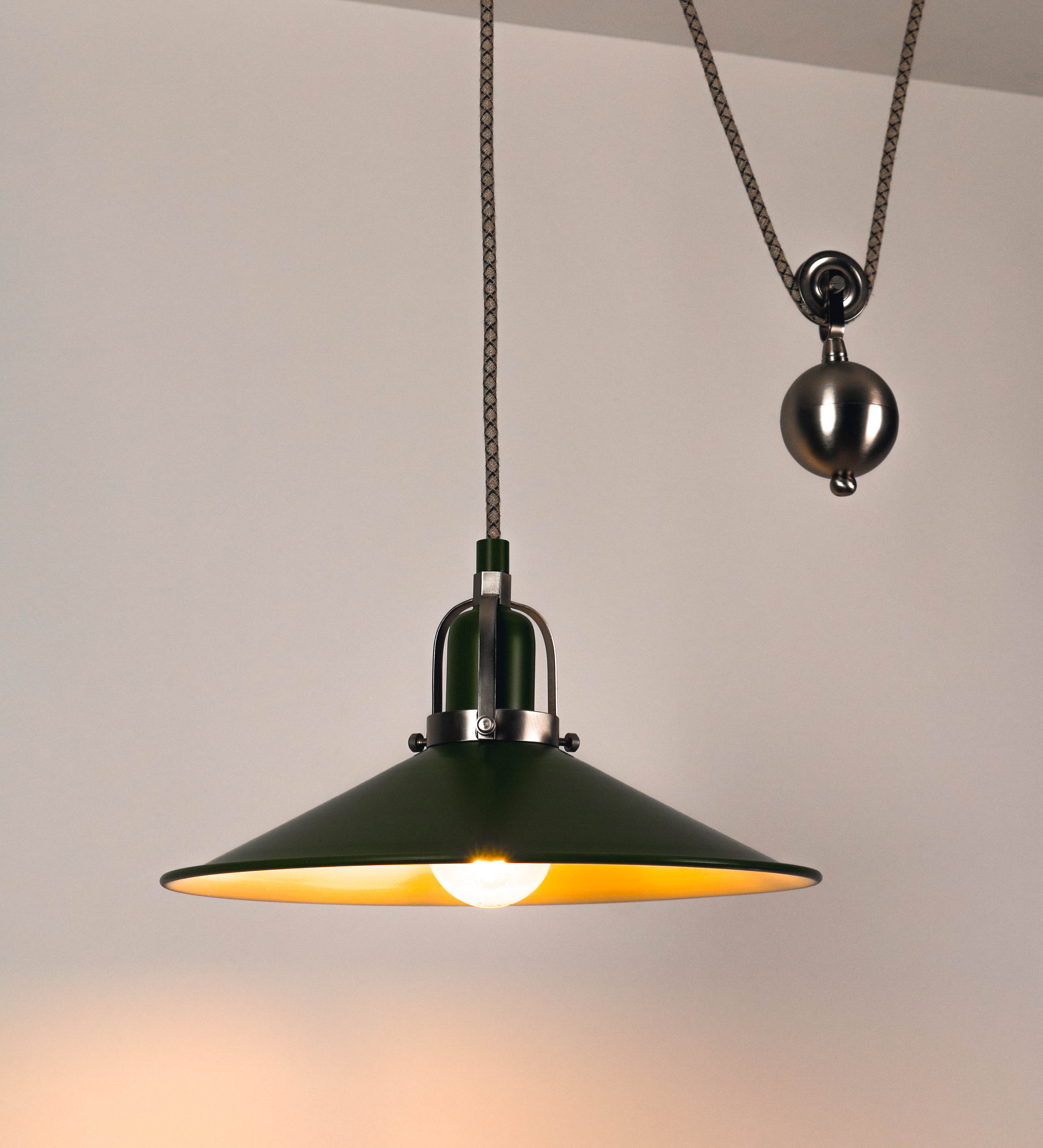 Suspensão de teto composta por uma base circular verde couve-de-bruxelas de onde desce um cabo com padrão de linhas castanhas e beges. O cabo passa por uma roldana em níquel escovado fixa ao teto, e desce até ao chapéu. Entre a base e a roldana o cabo sustenta uma esfera em níquel escovado, que atua como um contrapeso, para ajustar a altura. O chapéu é verde, de formato cónico invertido, uma borda levantada e interior branco. No topo do chapéu há um suporte em níquel escovado que conecta o chapéu ao cabo.