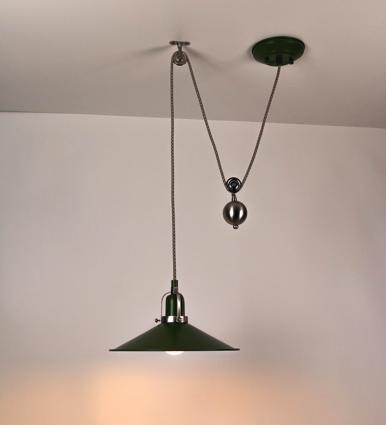 Suspensão de teto composta por uma base circular verde couve-de-bruxelas de onde desce um cabo com padrão de linhas castanhas e beges. O cabo passa por uma roldana em níquel escovado fixa ao teto, e desce até ao chapéu. Entre a base e a roldana o cabo sustenta uma esfera em níquel escovado, que atua como um contrapeso, para ajustar a altura. O chapéu é verde, de formato cónico invertido, uma borda levantada e interior branco. No topo do chapéu há um suporte em níquel escovado que conecta o chapéu ao cabo.