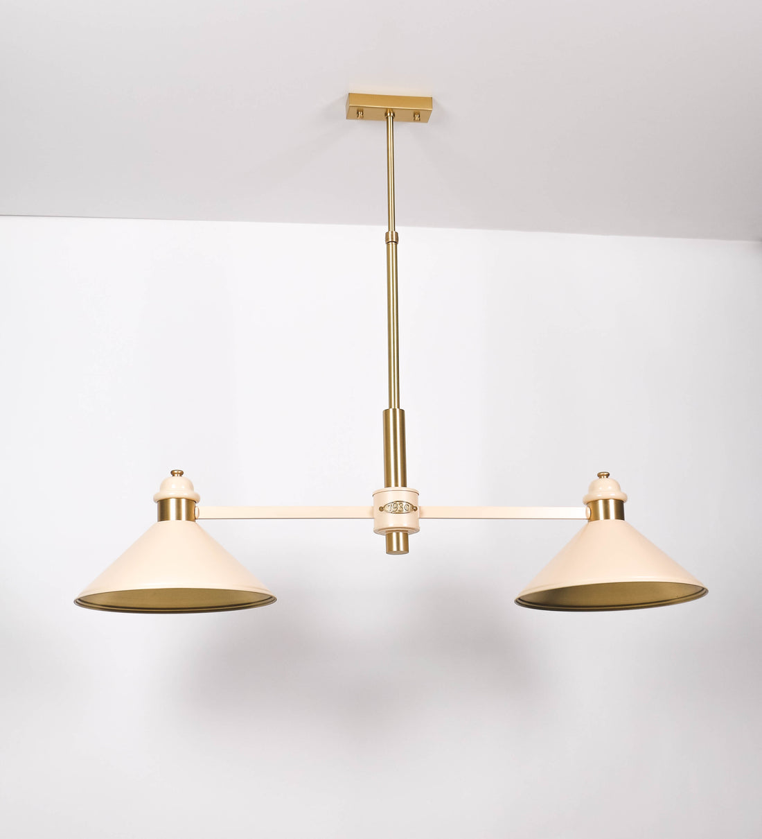 Suspensão de teto moderna e sofisticada. A suspensão é composta por uma base retangular em latão escovado, de onde desce um tubo cilíndrico até a estrutura principal e permite ajustar a altura da suspensão. A estrutura principal é composta por uma barra horizontal bege clara, que sustenta nas extremidades dois chapéus cónicos invertidos no mesmo tom, com interior em latão escovado. No centro da barra horizontal está uma pequena placa oval, em latão escovado, com o ano 1930 inscrito em relevo.