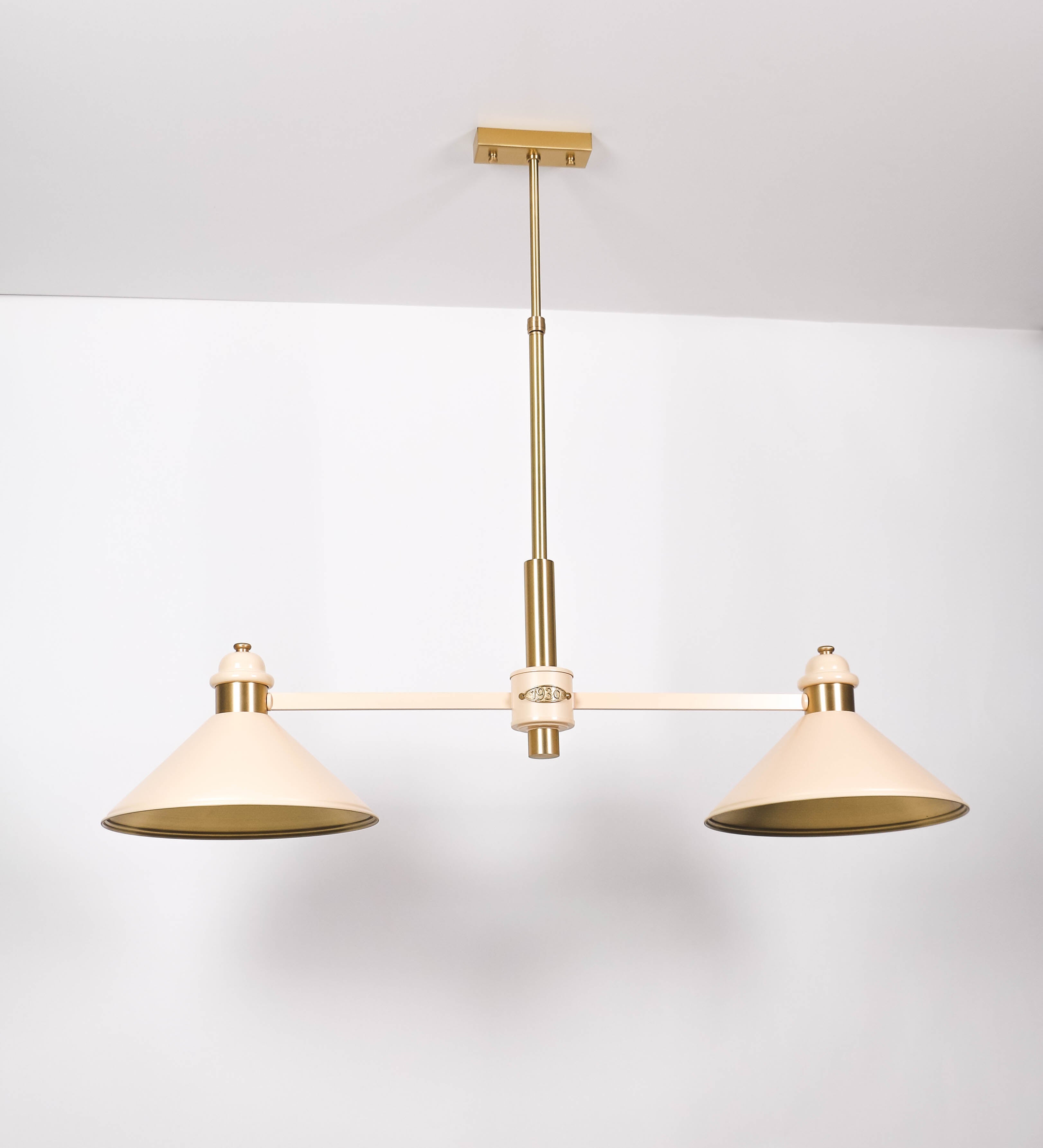 Suspensão de teto moderna e sofisticada. A suspensão é composta por uma base retangular em latão escovado, de onde desce um tubo cilíndrico até a estrutura principal e permite ajustar a altura da suspensão. A estrutura principal é composta por uma barra horizontal bege clara, que sustenta nas extremidades dois chapéus cónicos invertidos no mesmo tom, com interior em latão escovado. No centro da barra horizontal está uma pequena placa oval, em latão escovado, com o ano 1930 inscrito em relevo.