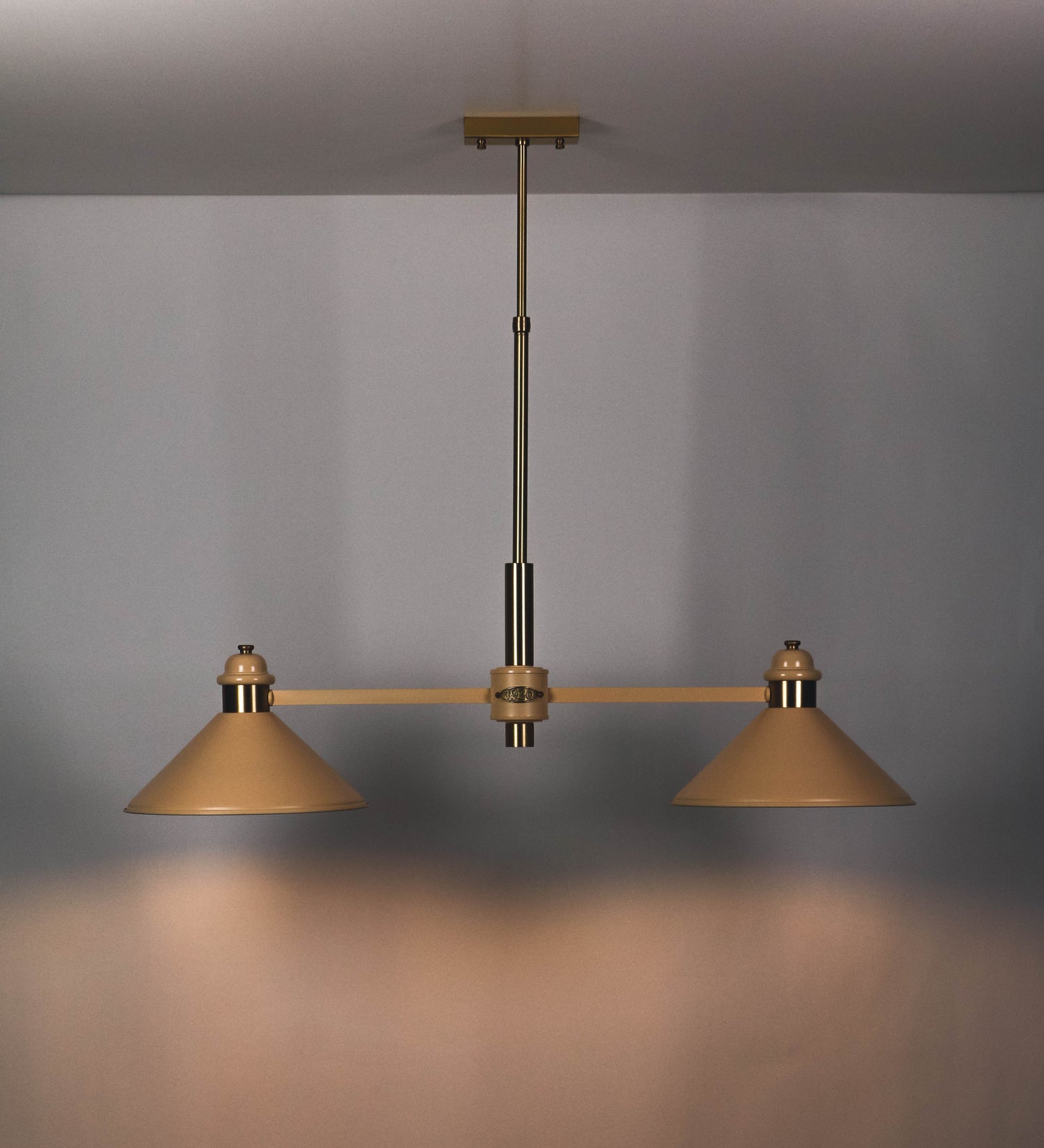 Suspensão de teto moderna e sofisticada. A suspensão é composta por uma base retangular em latão escovado, de onde desce um tubo cilíndrico até a estrutura principal e permite ajustar a altura da suspensão. A estrutura principal é composta por uma barra horizontal bege clara, que sustenta nas extremidades dois chapéus cónicos invertidos no mesmo tom, com interior em latão escovado. No centro da barra horizontal está uma pequena placa oval, em latão escovado, com o ano 1930 inscrito em relevo.