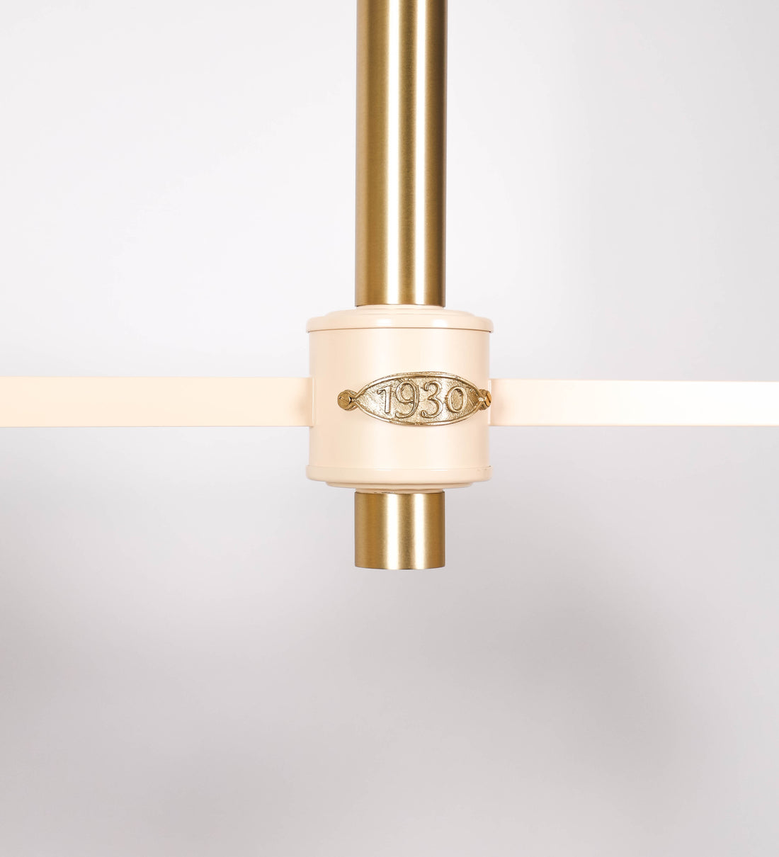 Suspensão de teto moderna e sofisticada. A suspensão é composta por uma base retangular em latão escovado, de onde desce um tubo cilíndrico até a estrutura principal e permite ajustar a altura da suspensão. A estrutura principal é composta por uma barra horizontal bege clara, que sustenta nas extremidades dois chapéus cónicos invertidos no mesmo tom, com interior em latão escovado. No centro da barra horizontal está uma pequena placa oval, em latão escovado, com o ano 1930 inscrito em relevo.