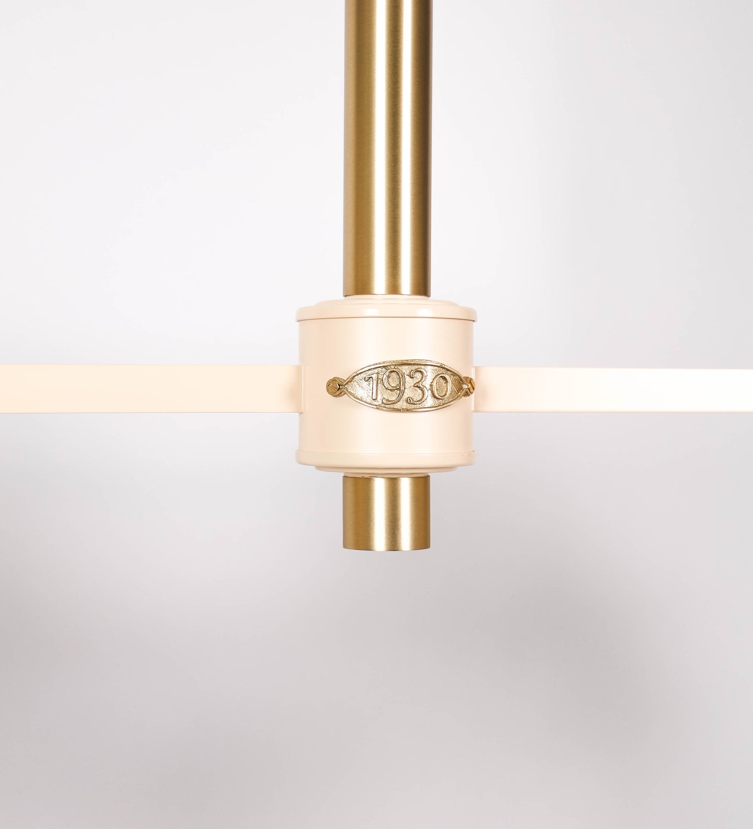 Suspensão de teto moderna e sofisticada. A suspensão é composta por uma base retangular em latão escovado, de onde desce um tubo cilíndrico até a estrutura principal e permite ajustar a altura da suspensão. A estrutura principal é composta por uma barra horizontal bege clara, que sustenta nas extremidades dois chapéus cónicos invertidos no mesmo tom, com interior em latão escovado. No centro da barra horizontal está uma pequena placa oval, em latão escovado, com o ano 1930 inscrito em relevo.