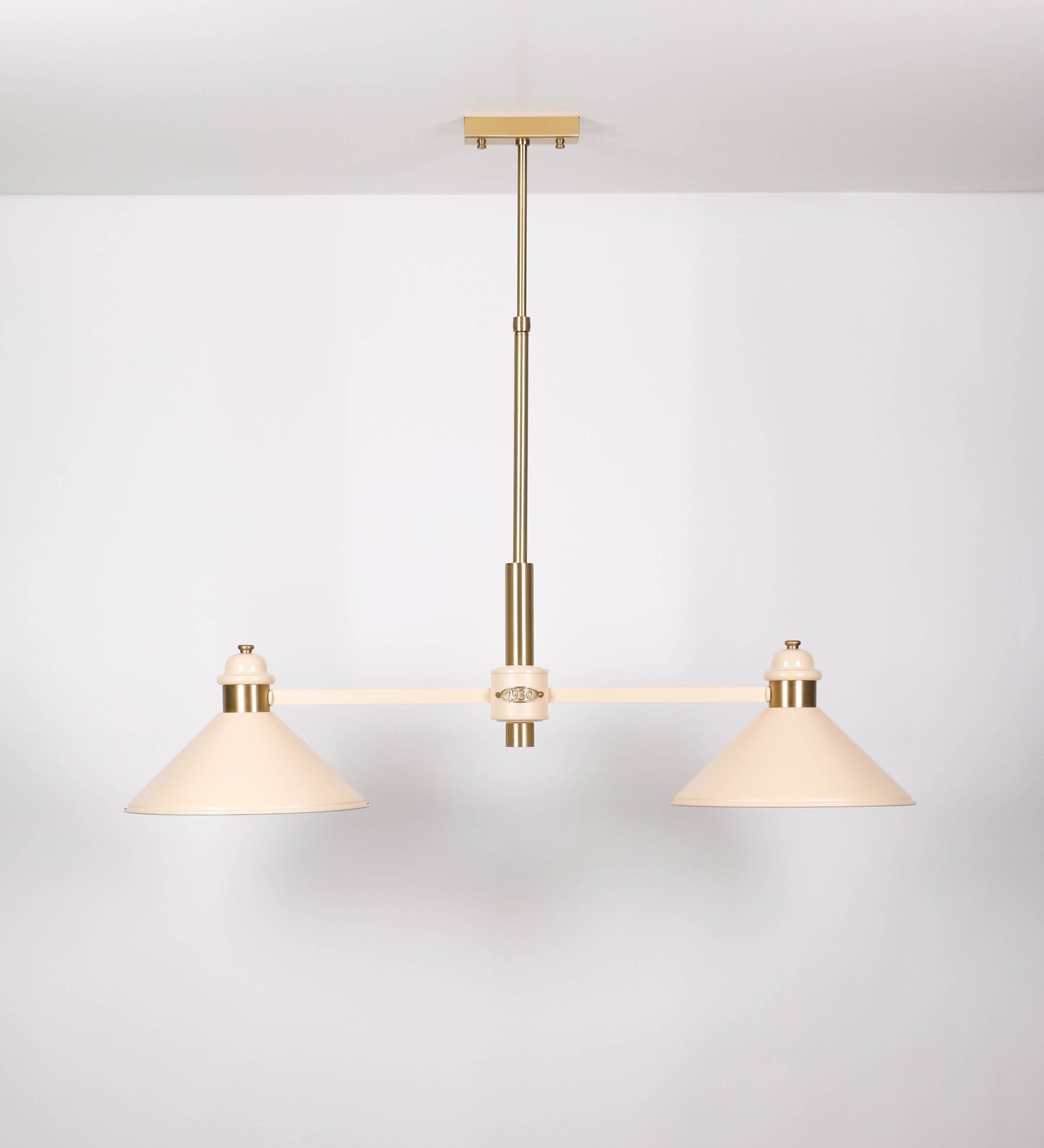 Suspensão de teto moderna e sofisticada. A suspensão é composta por uma base retangular em latão escovado, de onde desce um tubo cilíndrico até a estrutura principal e permite ajustar a altura da suspensão. A estrutura principal é composta por uma barra horizontal bege clara, que sustenta nas extremidades dois chapéus cónicos invertidos no mesmo tom, com interior em latão escovado. No centro da barra horizontal está uma pequena placa oval, em latão escovado, com o ano 1930 inscrito em relevo.