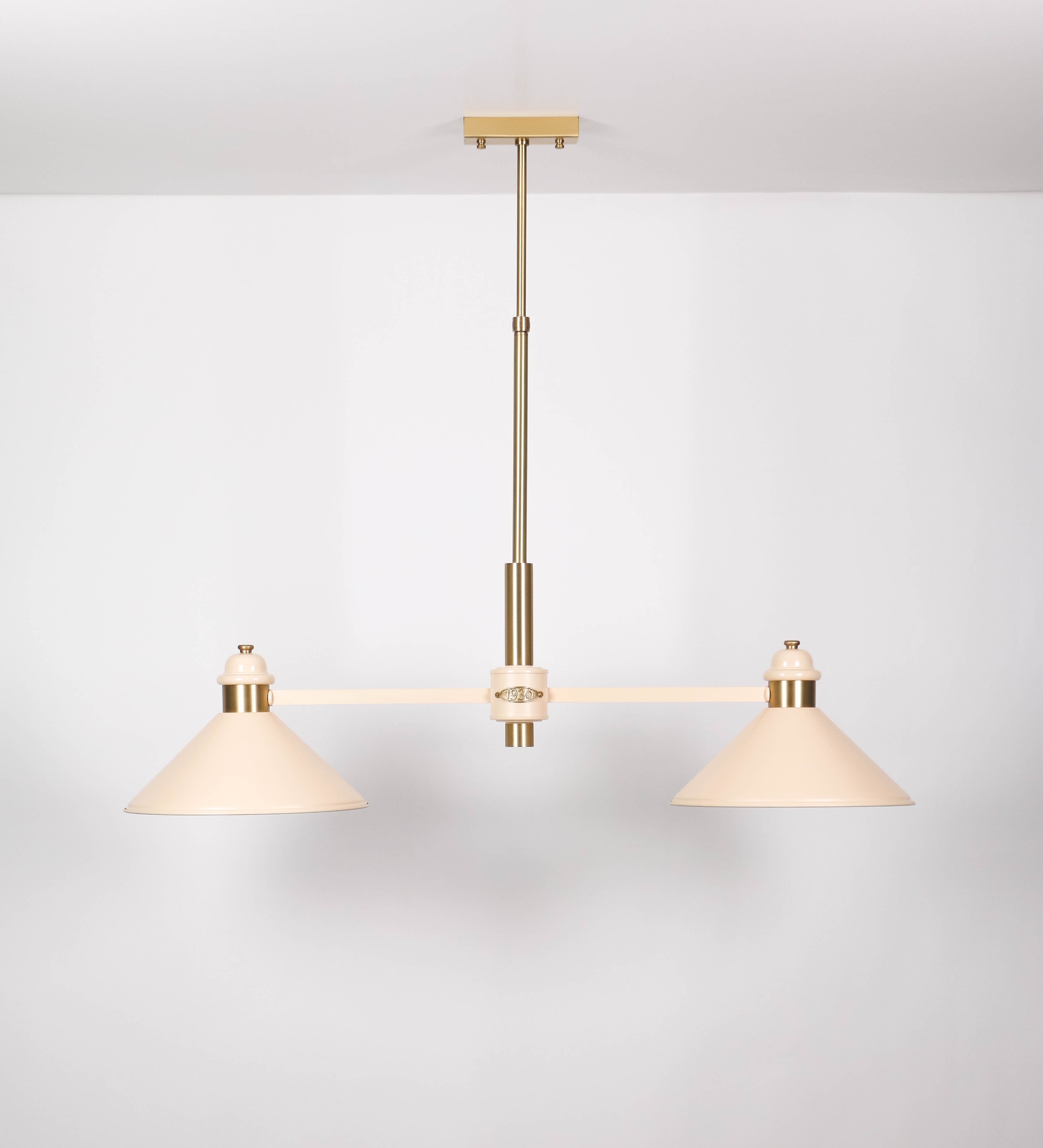 Suspensão de teto moderna e sofisticada. A suspensão é composta por uma base retangular em latão escovado, de onde desce um tubo cilíndrico até a estrutura principal e permite ajustar a altura da suspensão. A estrutura principal é composta por uma barra horizontal bege clara, que sustenta nas extremidades dois chapéus cónicos invertidos no mesmo tom, com interior em latão escovado. No centro da barra horizontal está uma pequena placa oval, em latão escovado, com o ano 1930 inscrito em relevo.